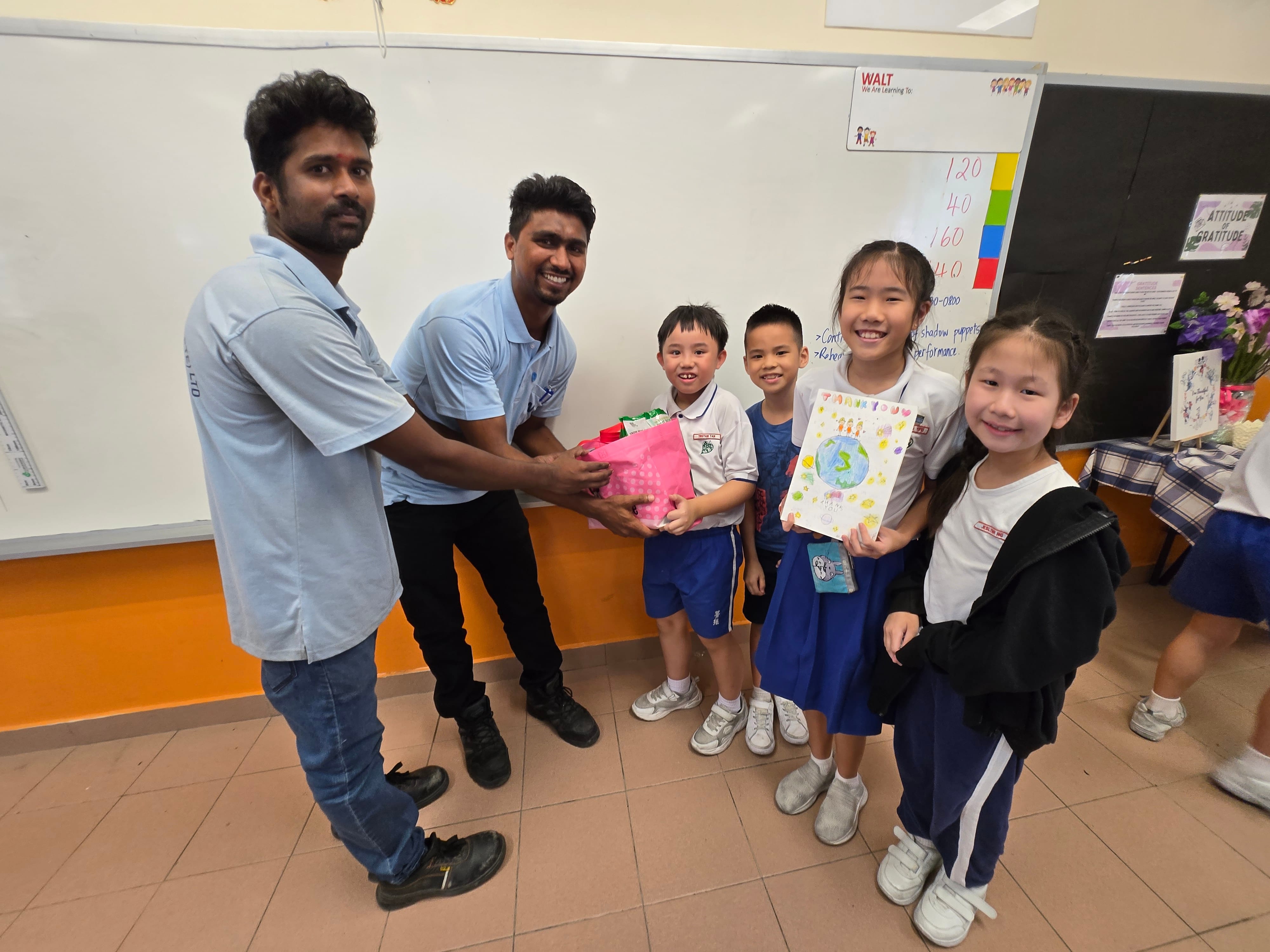 Two men receive a pink gift bag and a thank you card from four smiling children in a classroom.