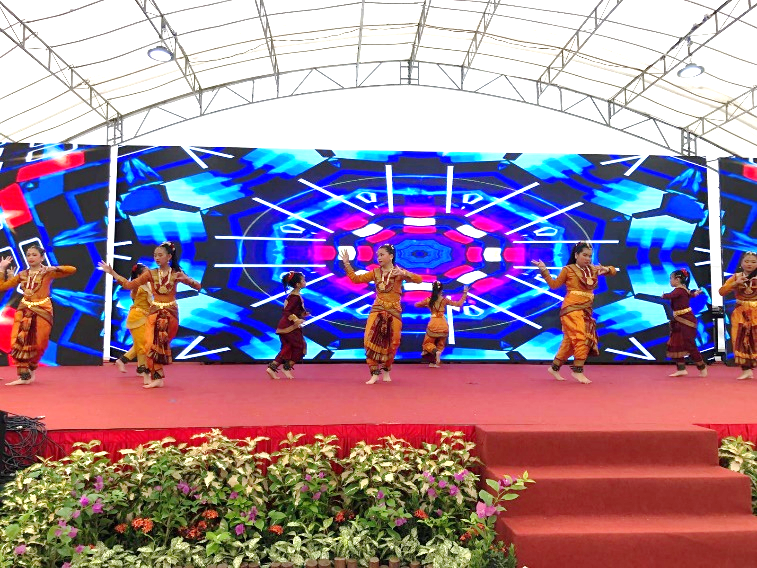 Girls in traditional orange costumes perform classical Indian dance on a red stage with a colorful LED backdrop.