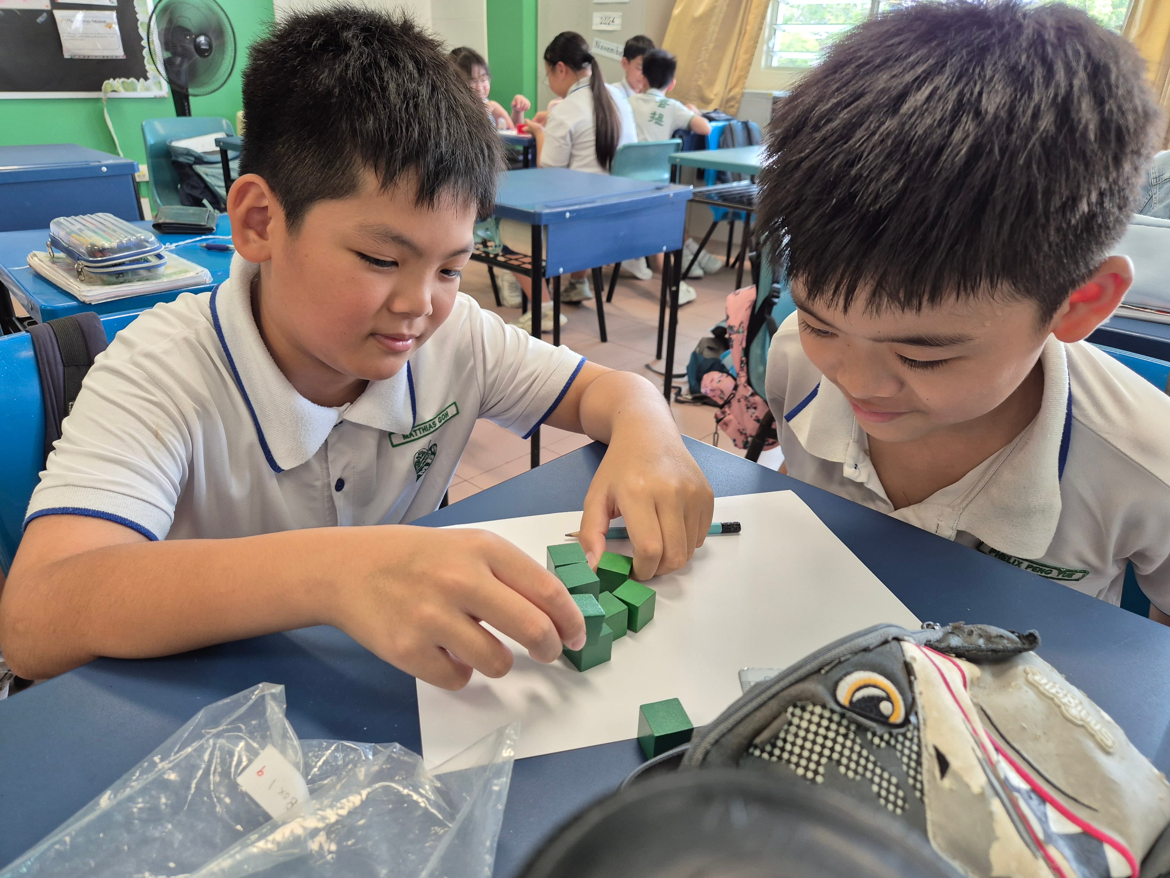 Two school boys in white uniforms arrange small green wooden blocks during Maths Learning session on a desk in a classroom.