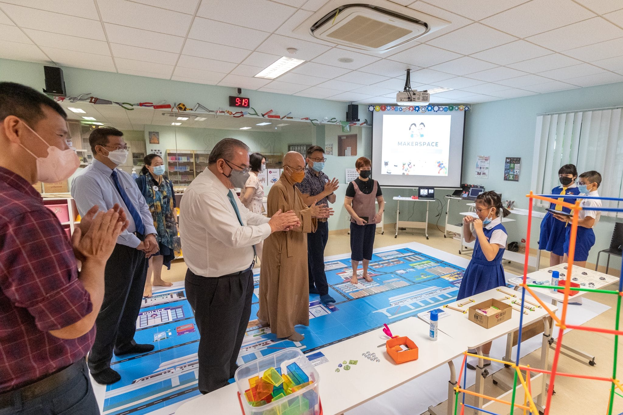 Adults and children in masks stand in a makerspace classroom with colorful building toys and a projected presentation.