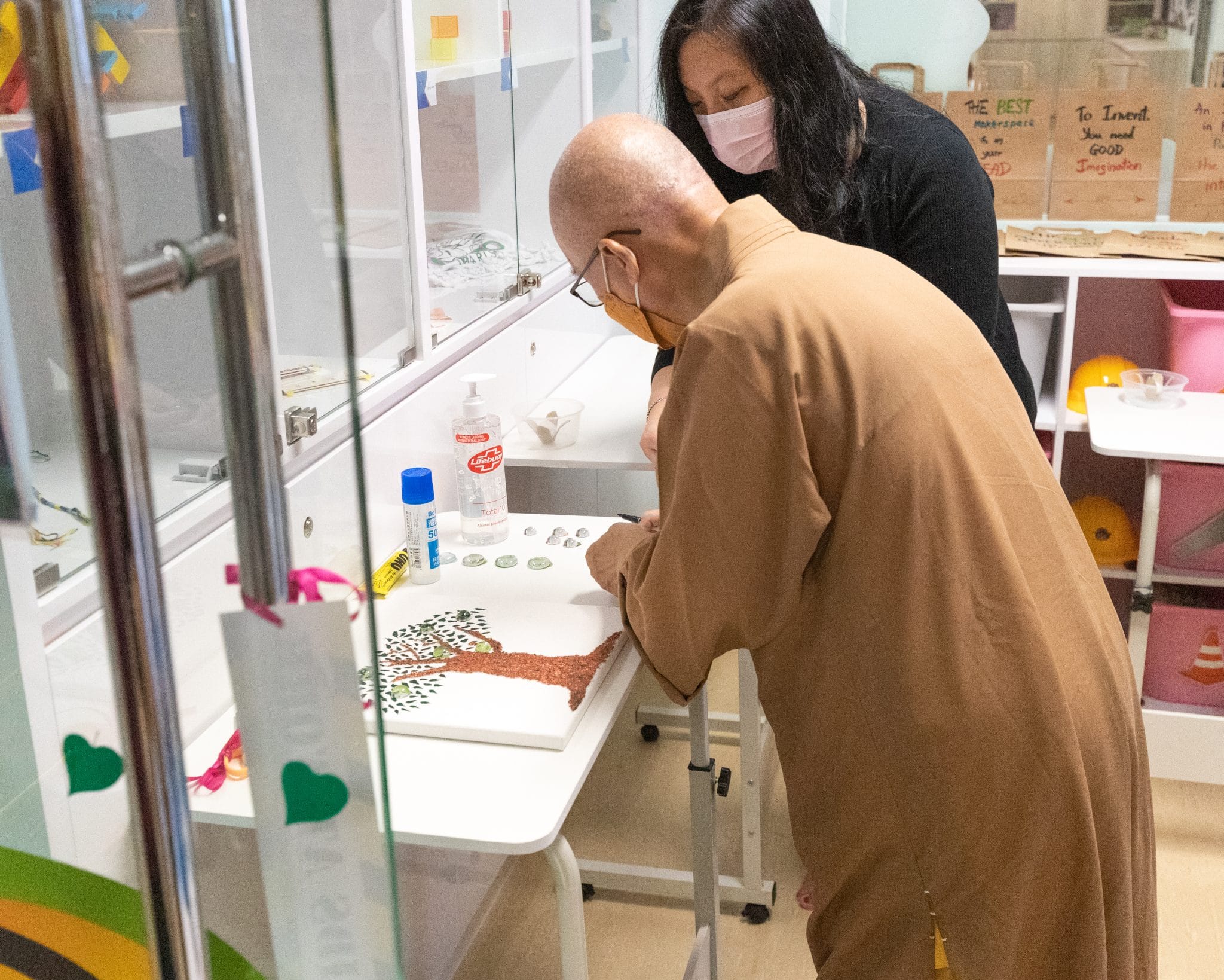 Two masked people lean over a table working on a craft project in a colorful indoor makerspace room.