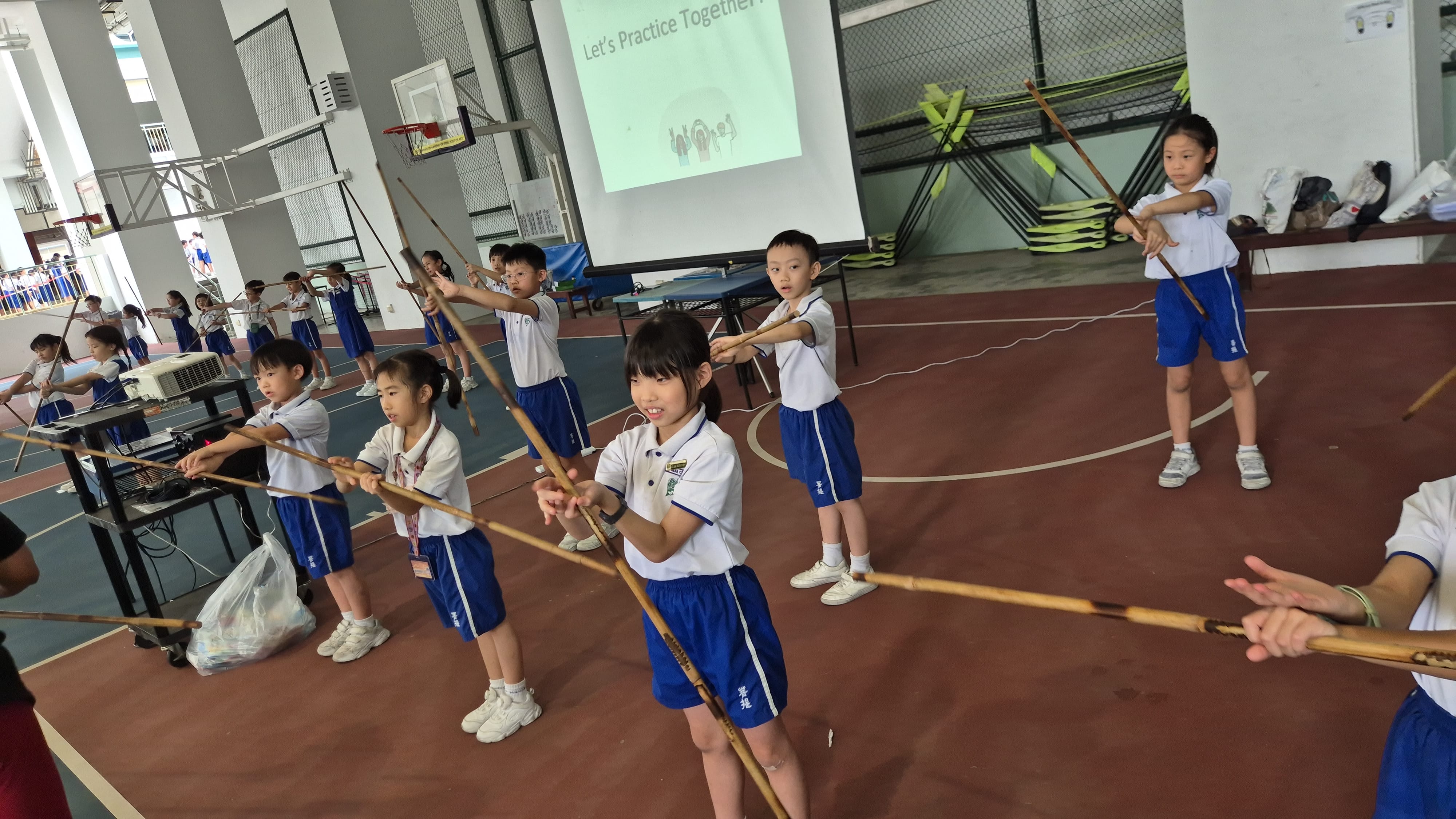 Young children in white and blue uniforms practicing archery with wooden bows in an indoor gymnasium.