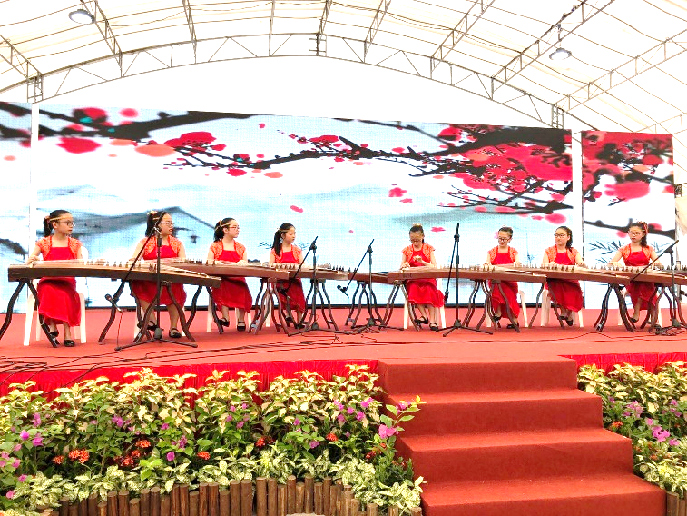 Girls in red dresses play guzheng on a decorated stage with a red blossom backdrop.