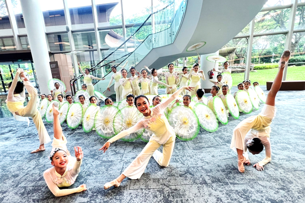 Group of young dancers in white and green outfits performing with decorative parasols in a modern building atrium.