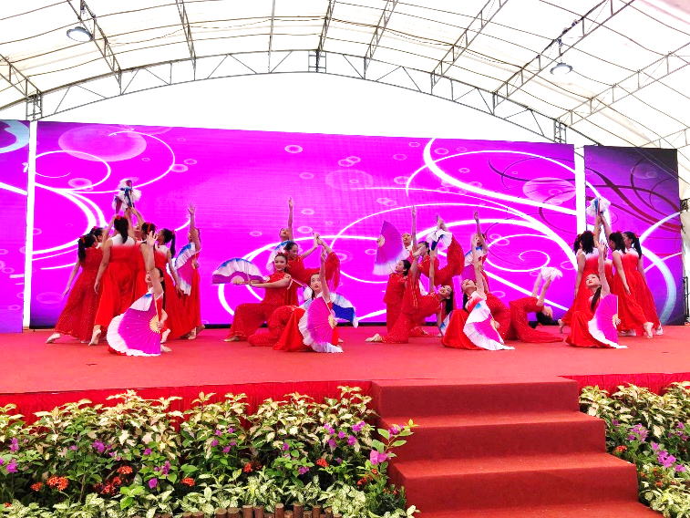 Group of women in red dresses performing a fan dance on a stage with a bright pink LED backdrop.