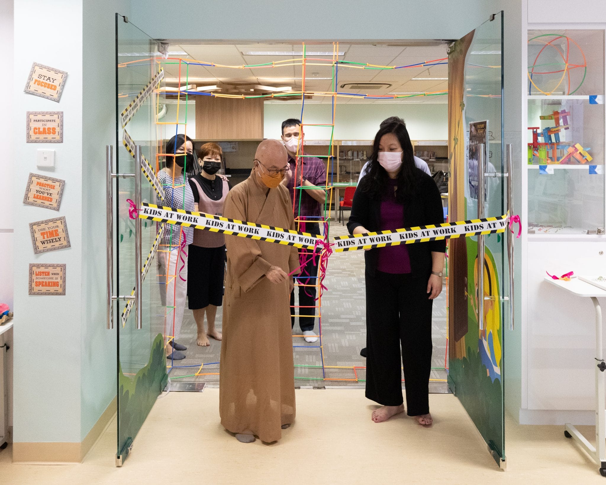Two masked people cut a Kids at Work ribbon at a colorful classroom entrance while others watch behind them.