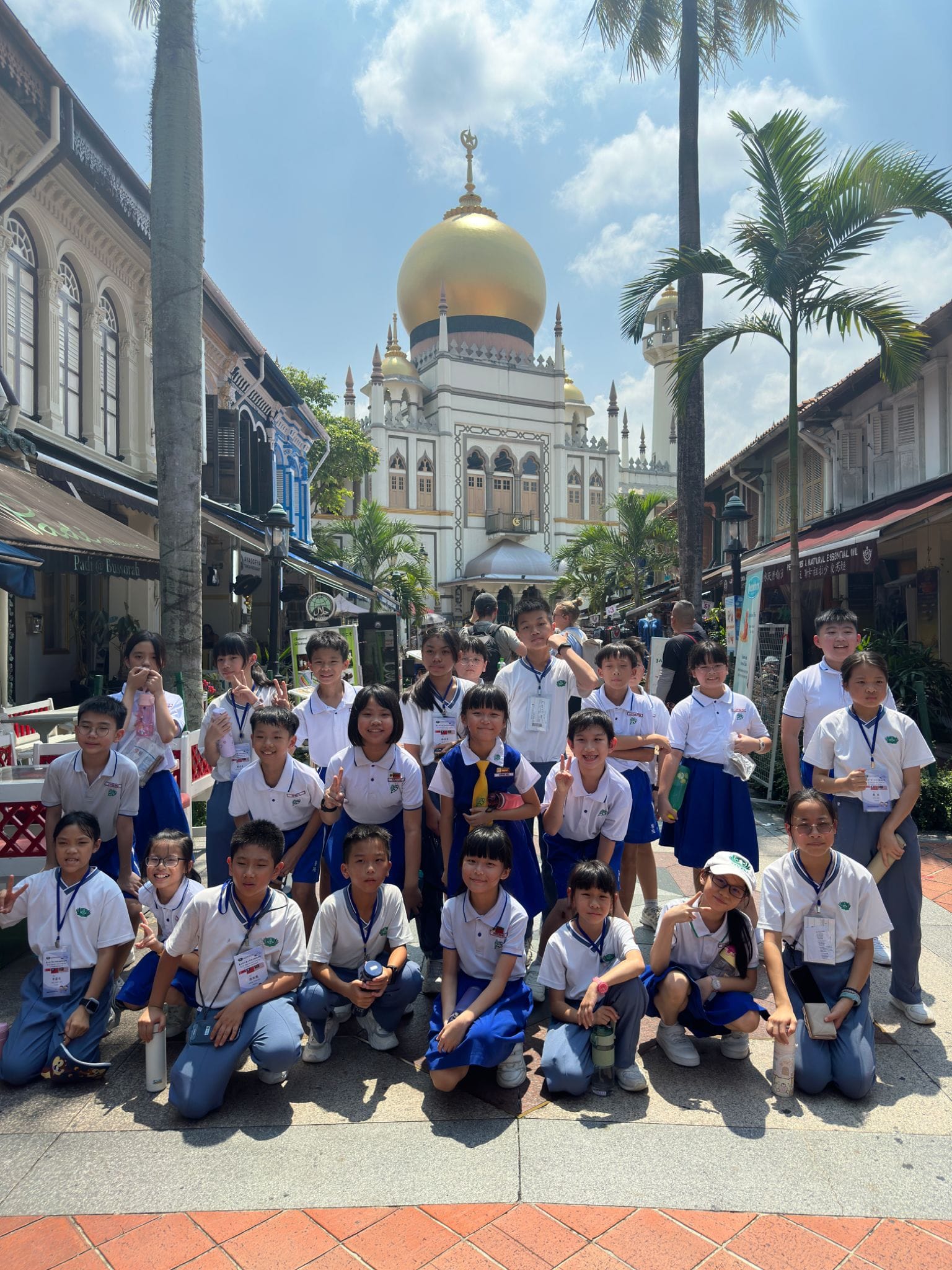 A group of children in school uniforms pose in front of a white mosque with a large golden dome.