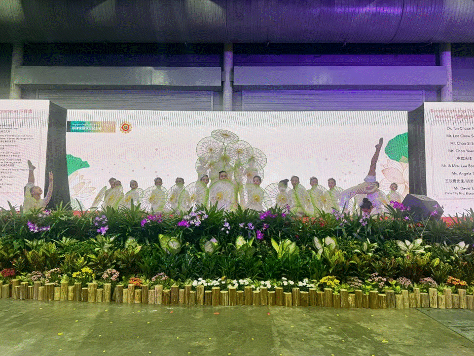 Dancers in white costumes holding parasols perform on a stage decorated with colorful flowers and green plants.
