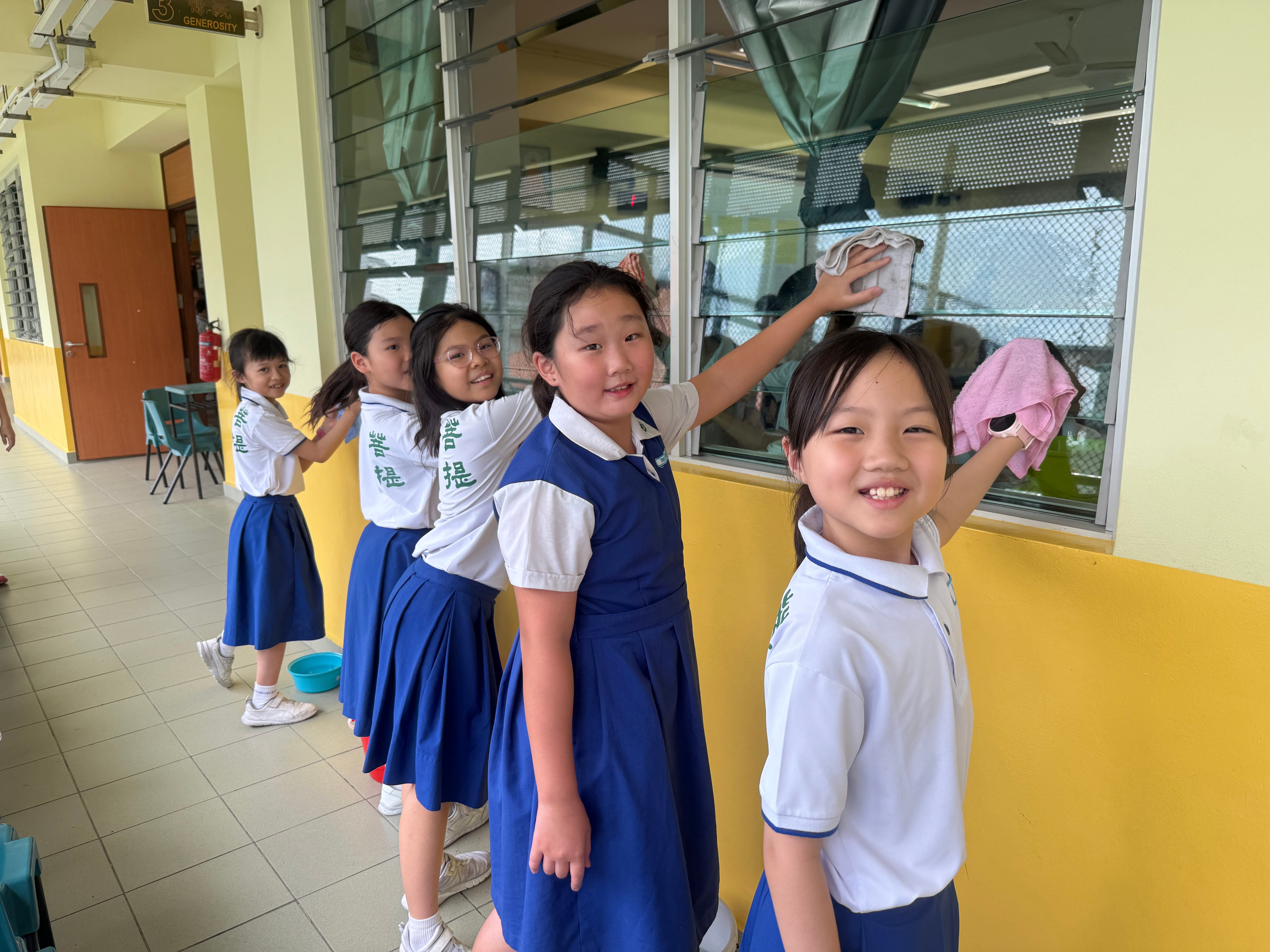 Five schoolgirls in blue and white uniforms smile while wiping windows with cloths in a school corridor.