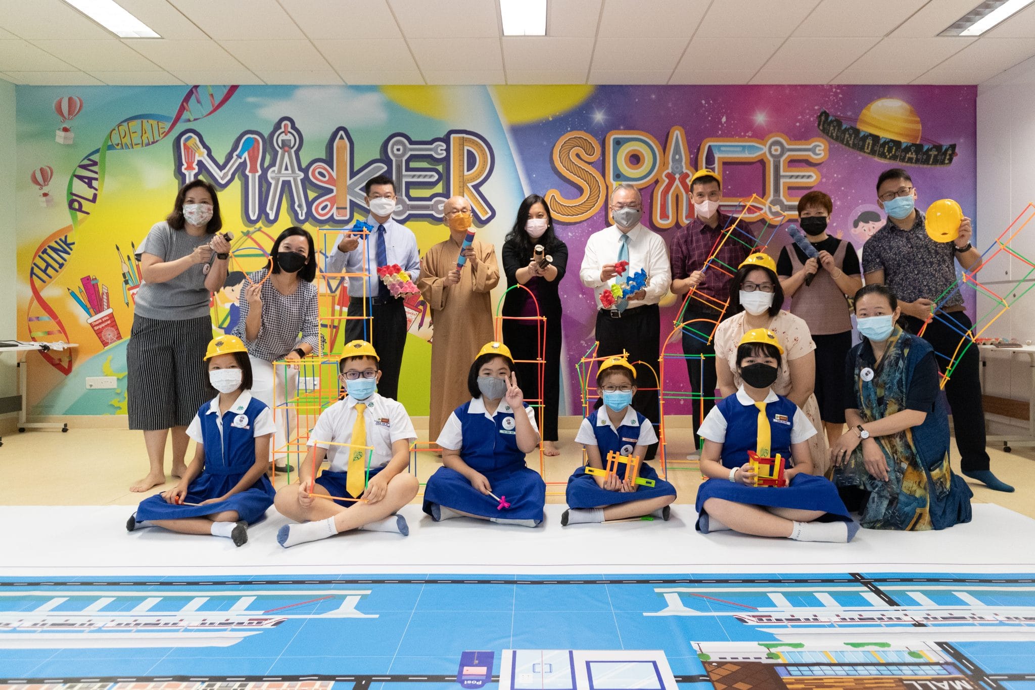 Adults and masked children in school uniforms pose with colorful building toys in a Maker Space room.