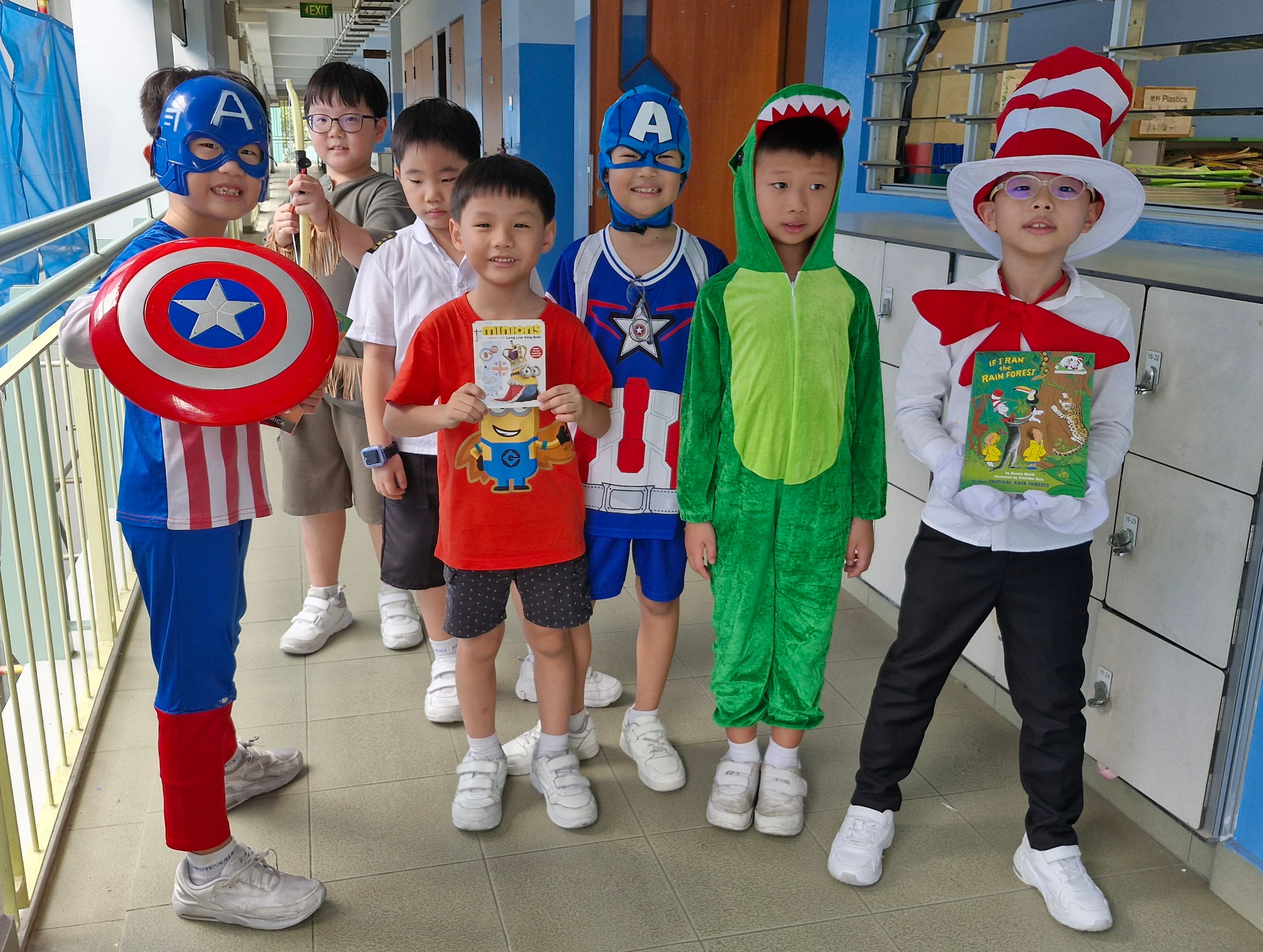 Six children in costumes including Captain America, a dinosaur, and Cat in the Hat pose outside their classroom.