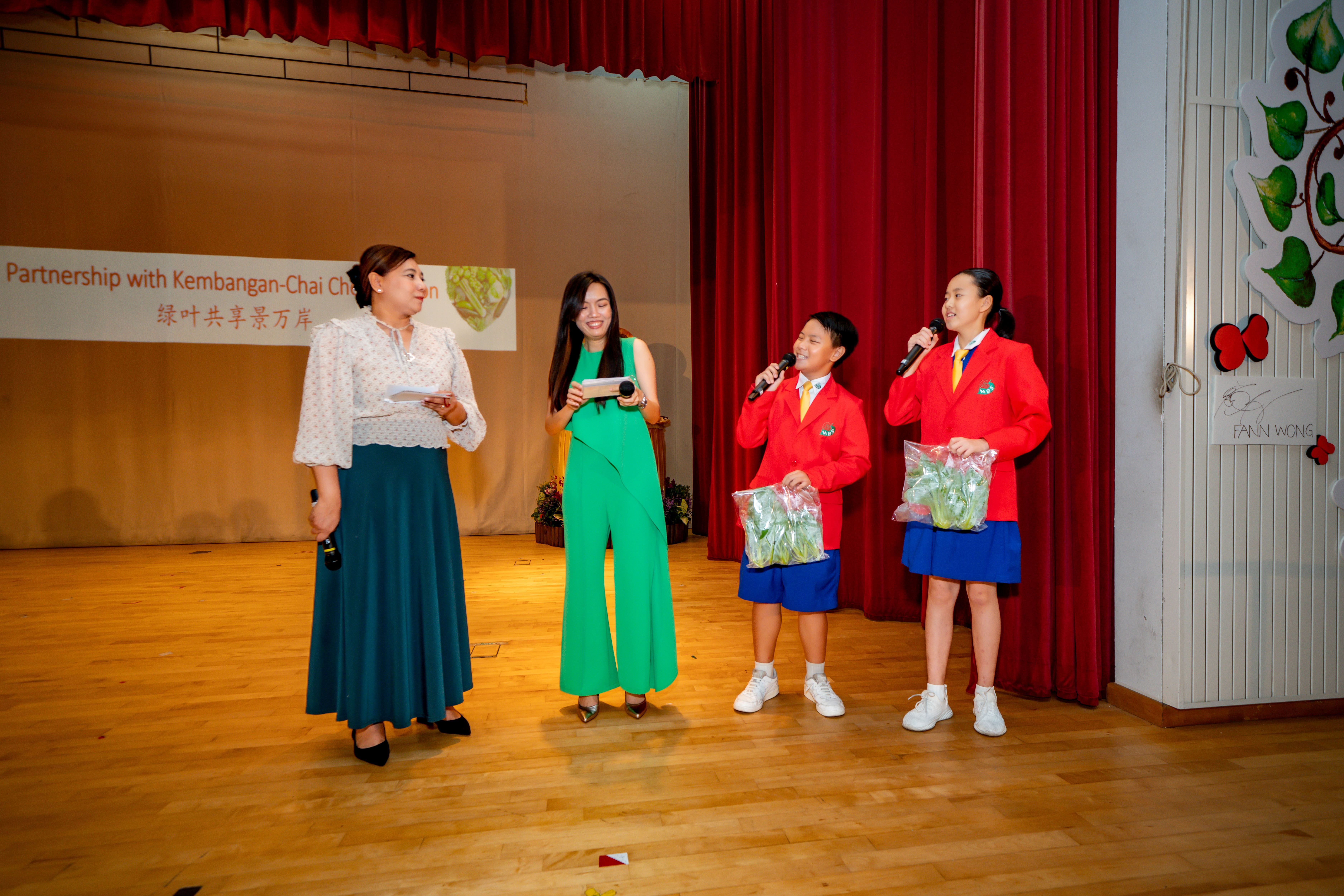 Two women and two children in red blazers speak into microphones on a school stage, holding bags of vegetables.