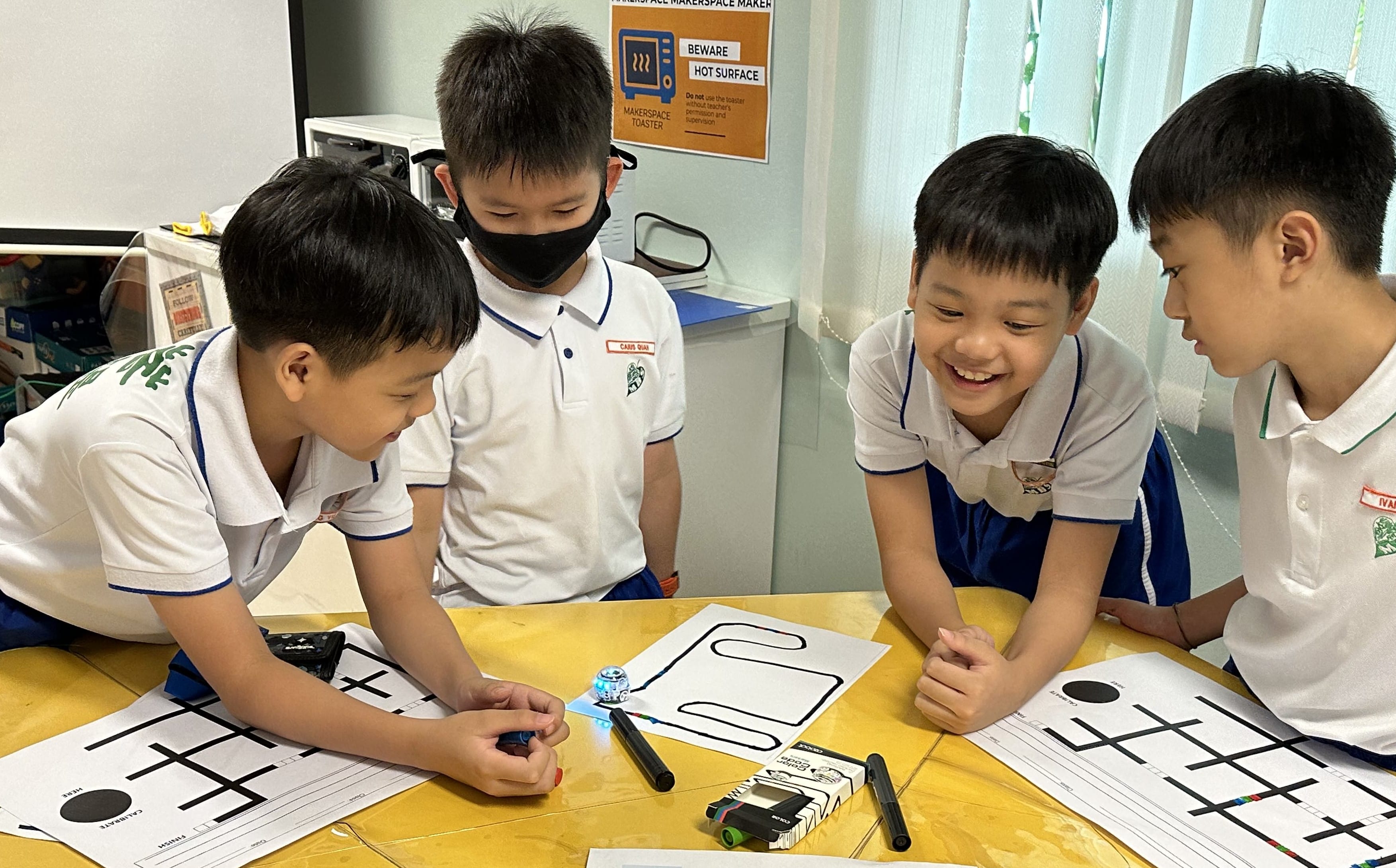 Four young students in school uniforms gather around a table with a small robot and printed track sheets.