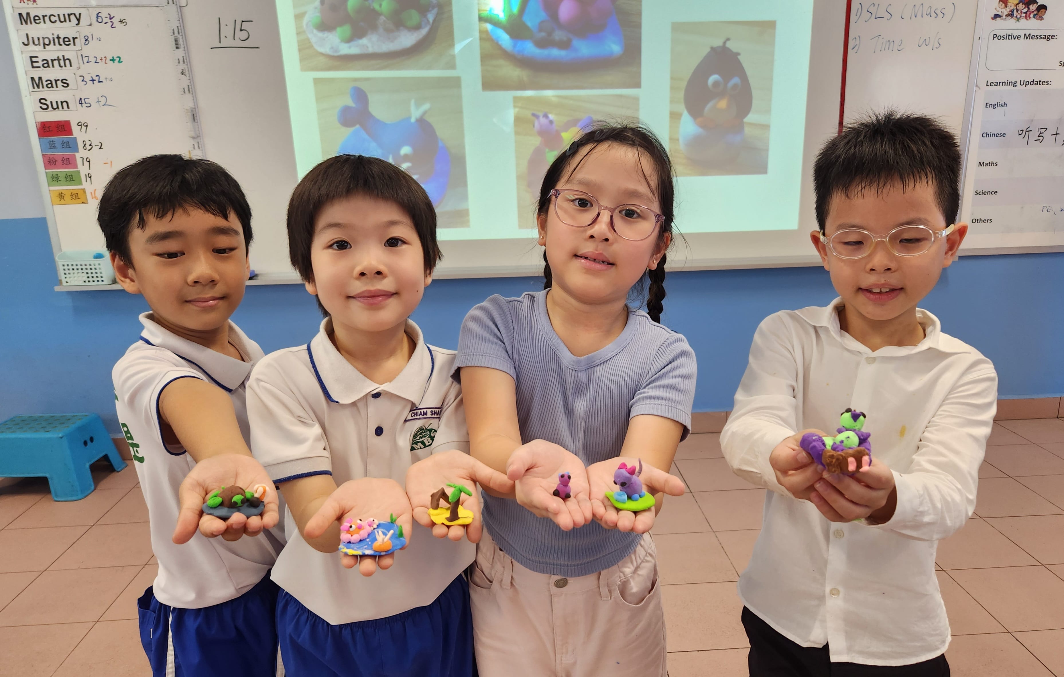 Four young students in a classroom holding out small colorful clay figurines they made, with a projector screen behind them.