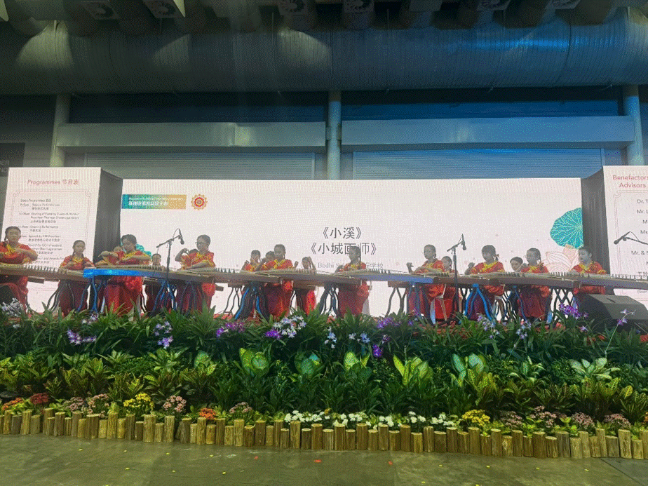 A group of performers in red outfits playing guzheng instruments on a decorated stage with flowers and Chinese text backdrop.
