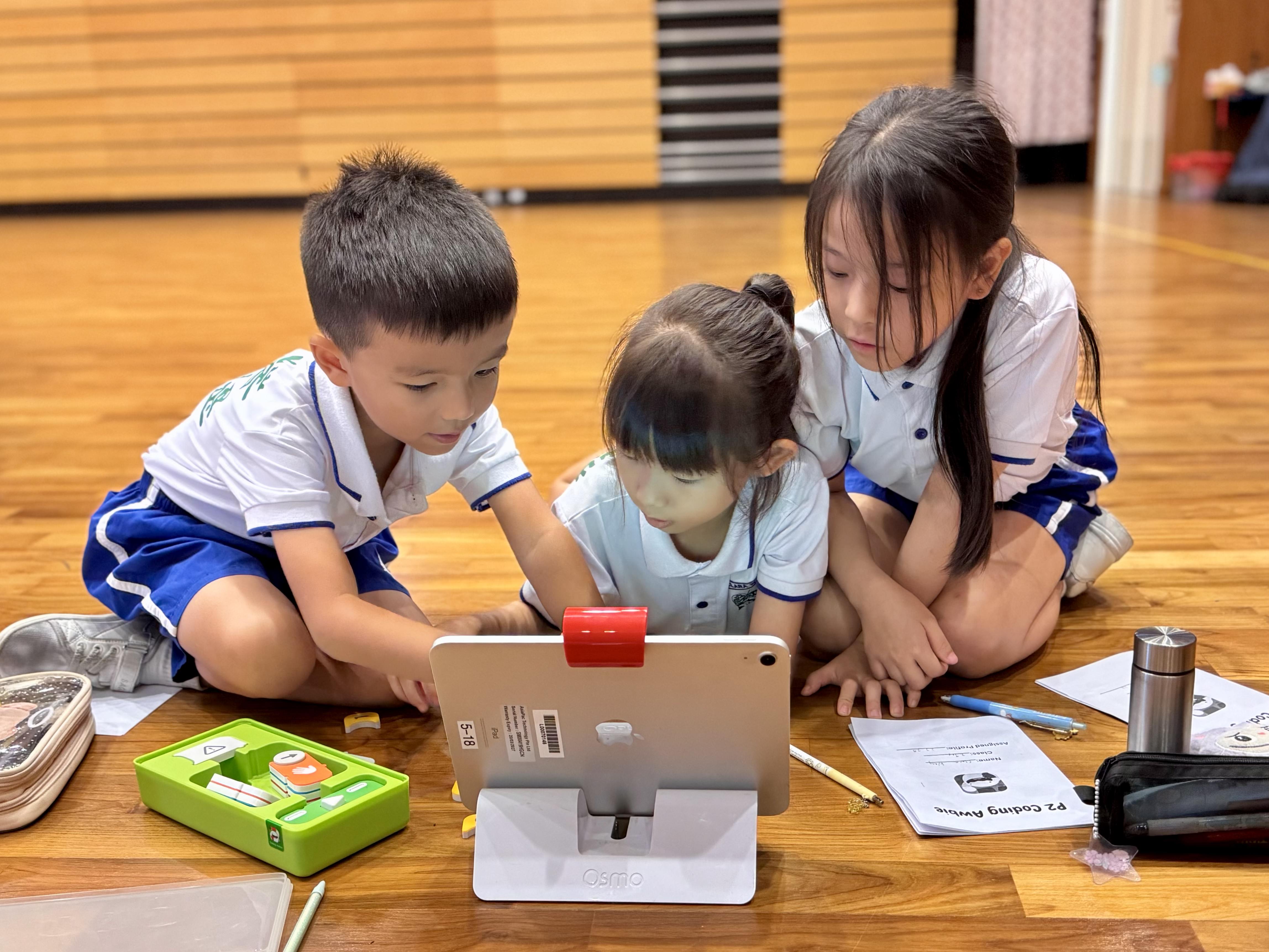 Three young children in school uniforms sit on a wooden floor, focused on a tablet in an Osmo stand.