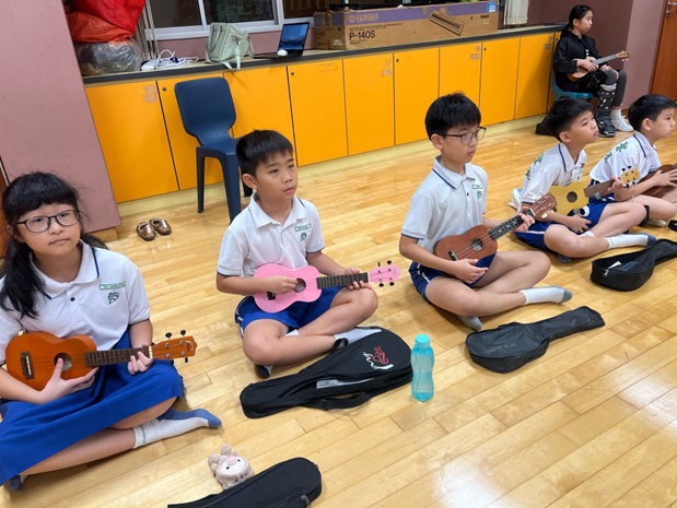 Several children in school uniforms sit cross-legged on a wooden floor holding colorful ukuleles during a music class.
