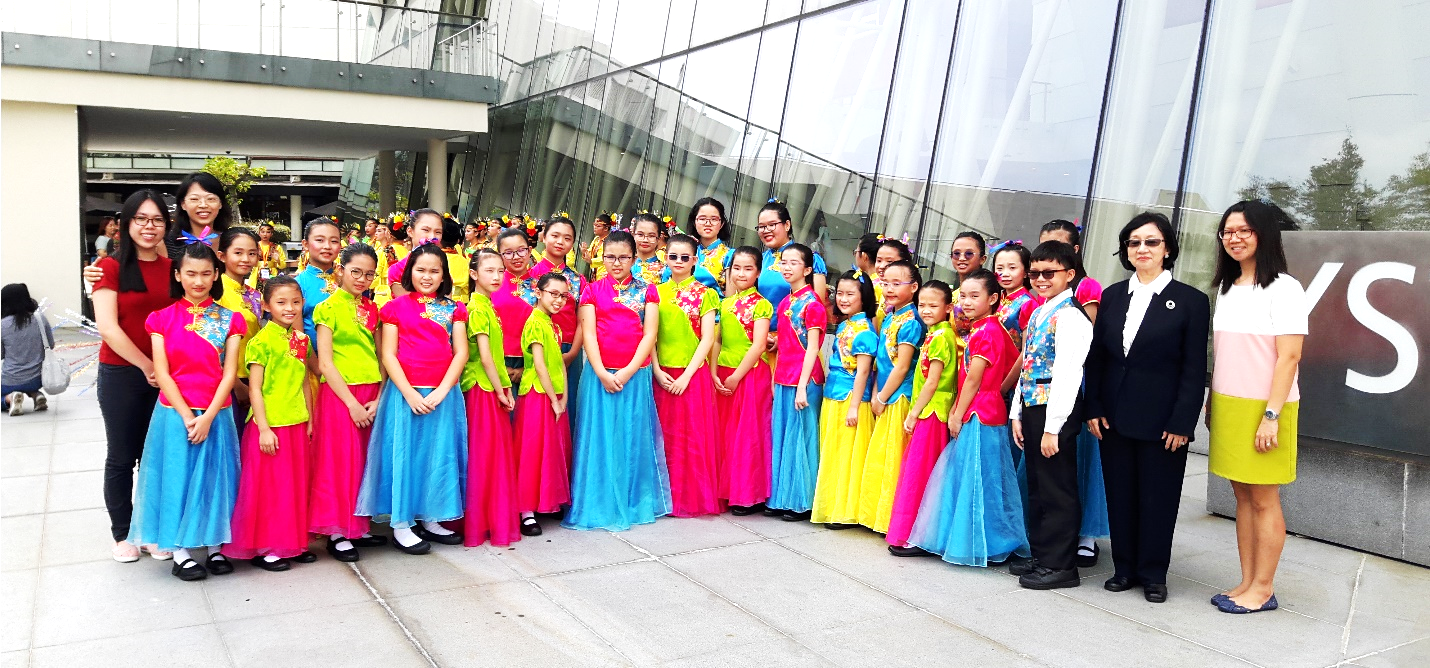 A large group of girls in colorful traditional Chinese costumes pose outdoors with two adult women near a glass building.