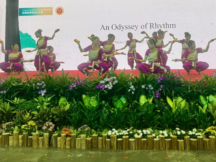 Dancers in purple costumes perform on stage behind a floral display, under a banner reading "An Odyssey of Rhythm."