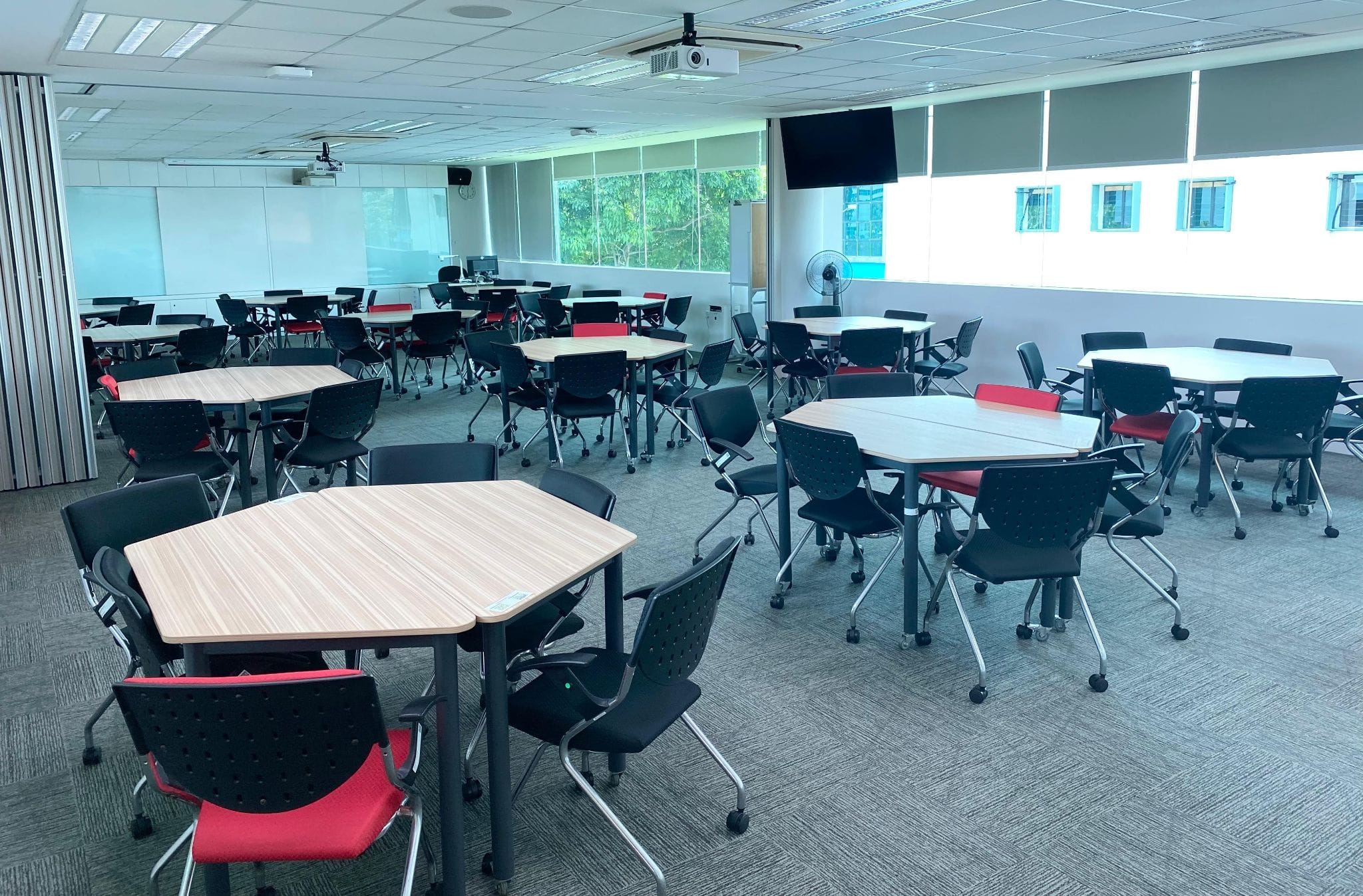 Empty classroom with clustered wooden tables, black and red chairs, a ceiling projector, and a wall-mounted TV screen.