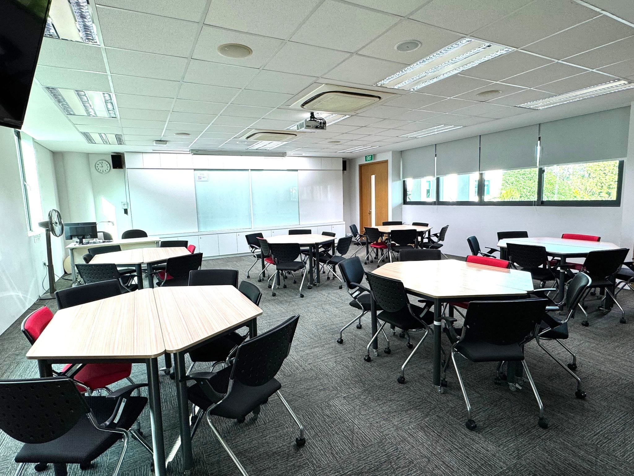 Empty classroom with hexagonal wooden tables, black and red chairs, a whiteboard, projector, and windows with blinds.