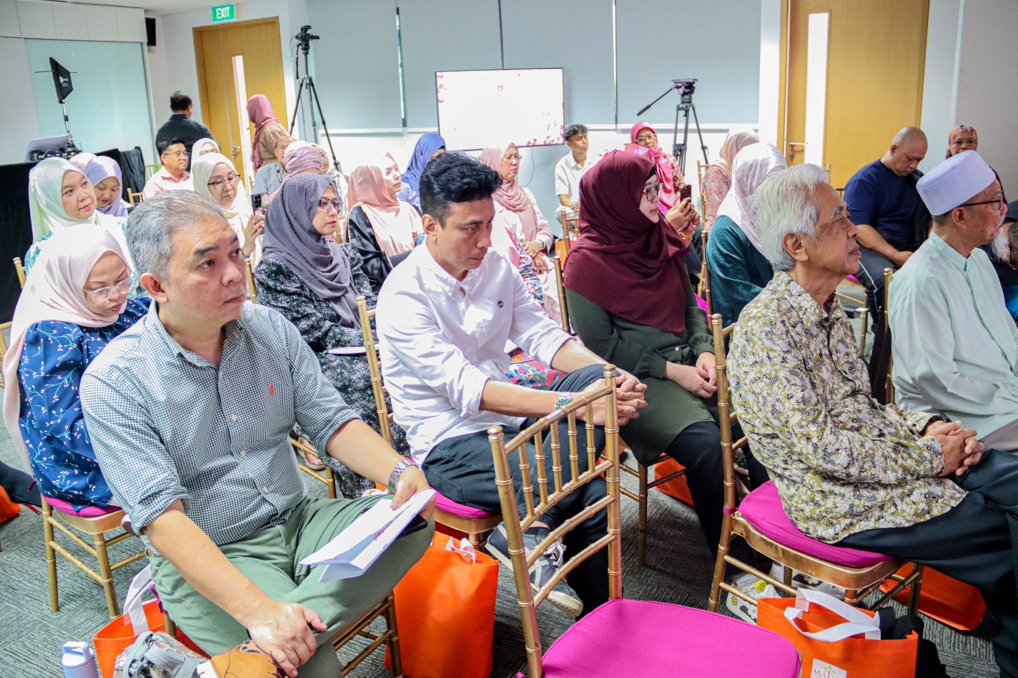 A crowd of people seated on gold chairs with pink cushions at an indoor event, many women wearing headscarves.