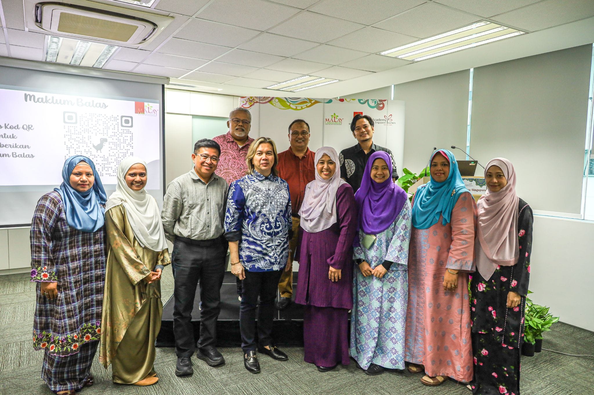A group of eleven people posing together in a conference room with a presentation screen showing a QR code behind them.