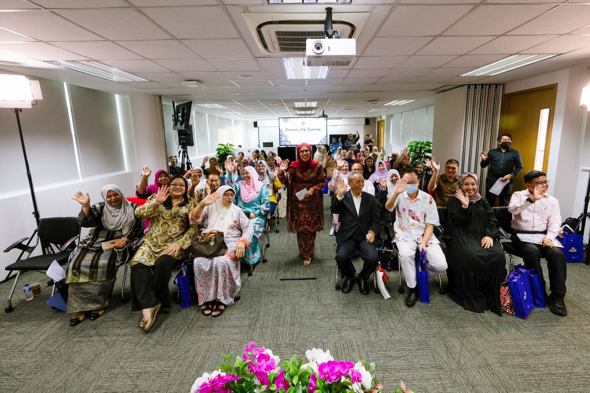 A large group of people in colorful attire wave and smile in a conference room with a projector screen behind them.