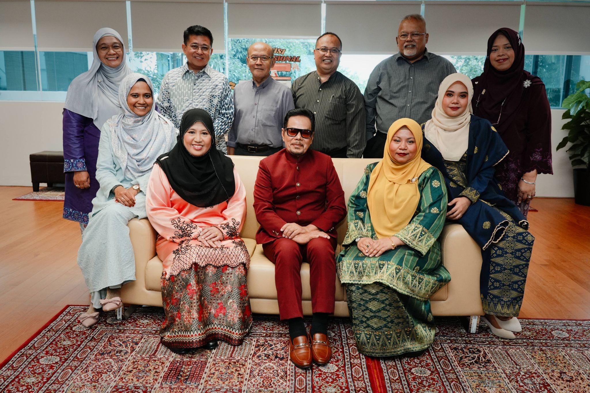 Group of eleven people in traditional Malay attire posing for a photo, seated and standing in an indoor setting.