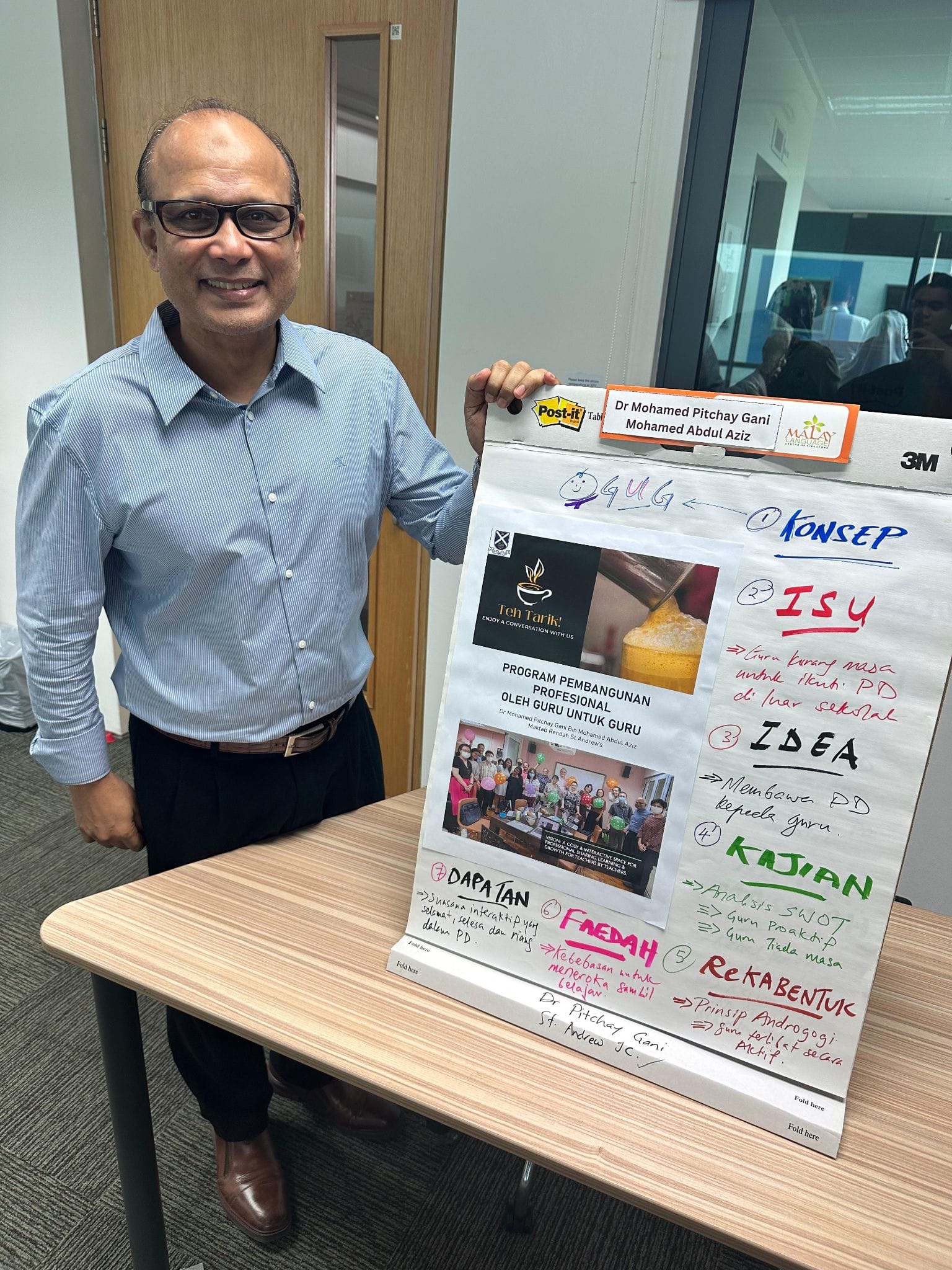 A smiling man in a blue shirt stands beside a flipchart with Malay text and colorful handwritten notes.