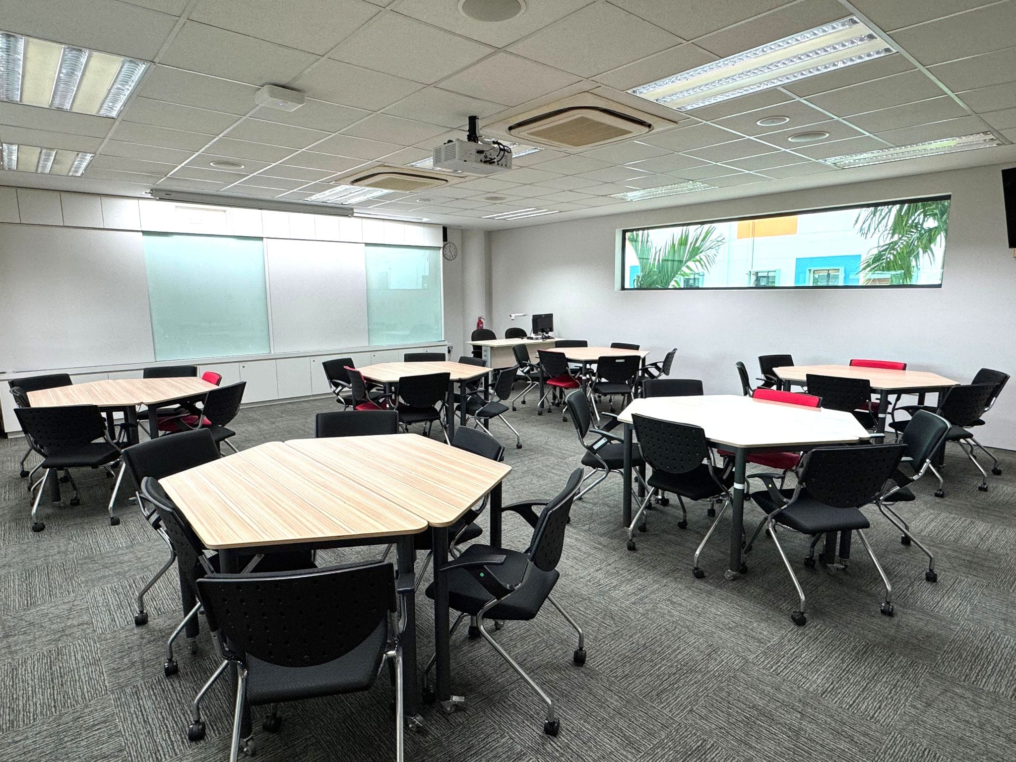 Empty classroom with hexagonal wooden tables, black rolling chairs, a ceiling projector, whiteboard, and a window with plants outside.