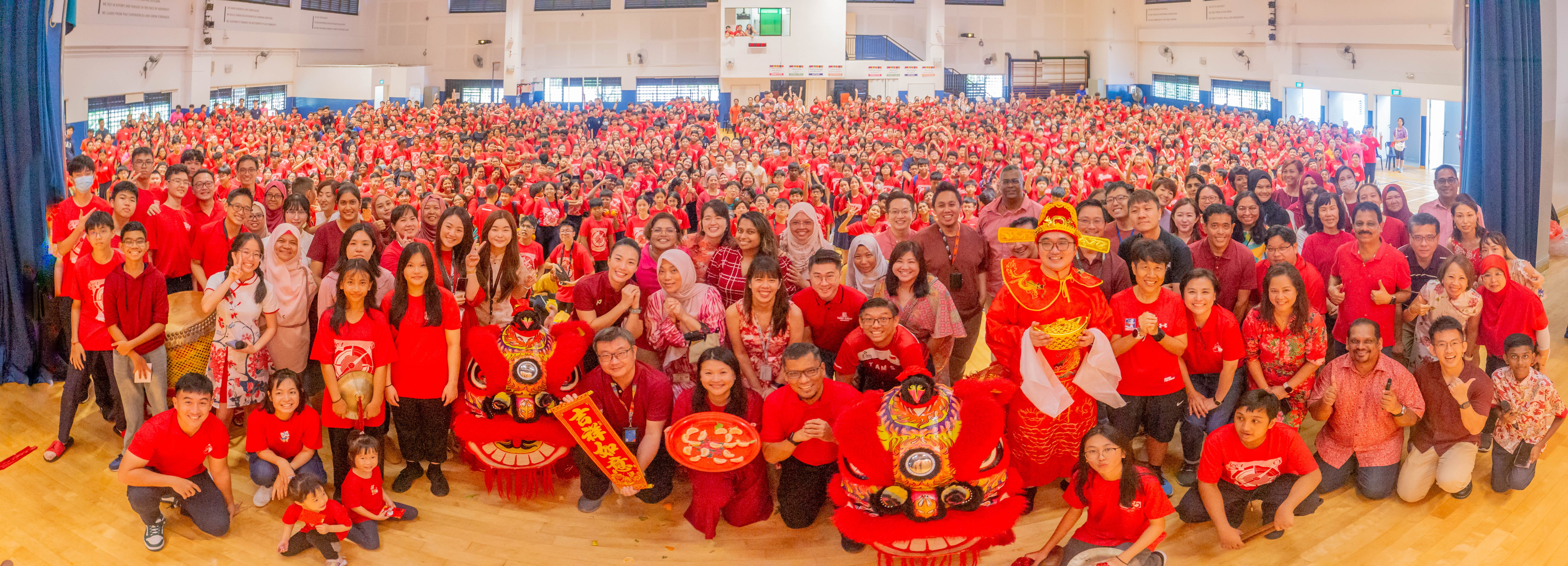 Huge crowd in a hall wearing red, posing with two lion dance heads and a person in festive red costume.