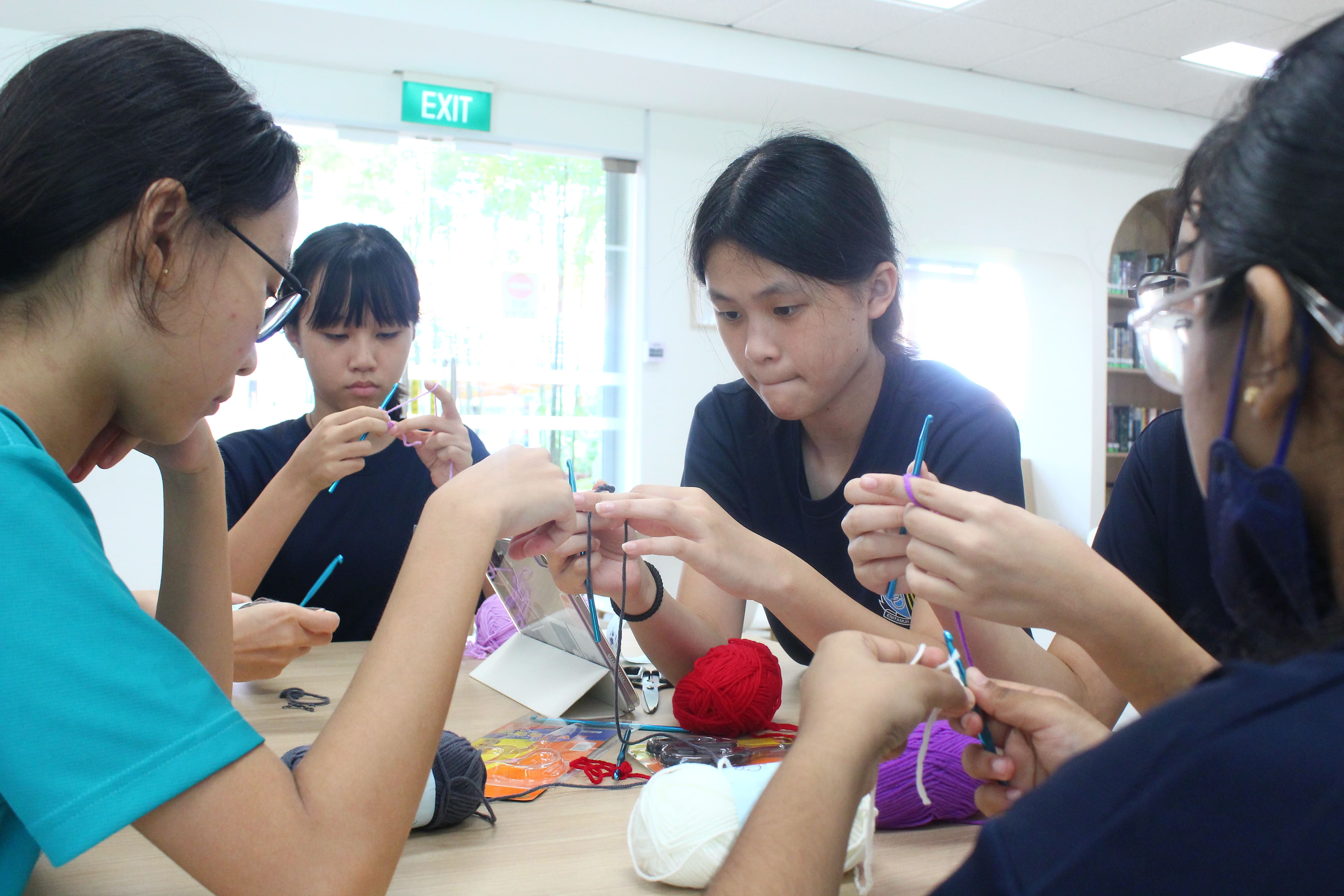 Four young students sit around a table crocheting with colorful yarn and hooks in a bright room.