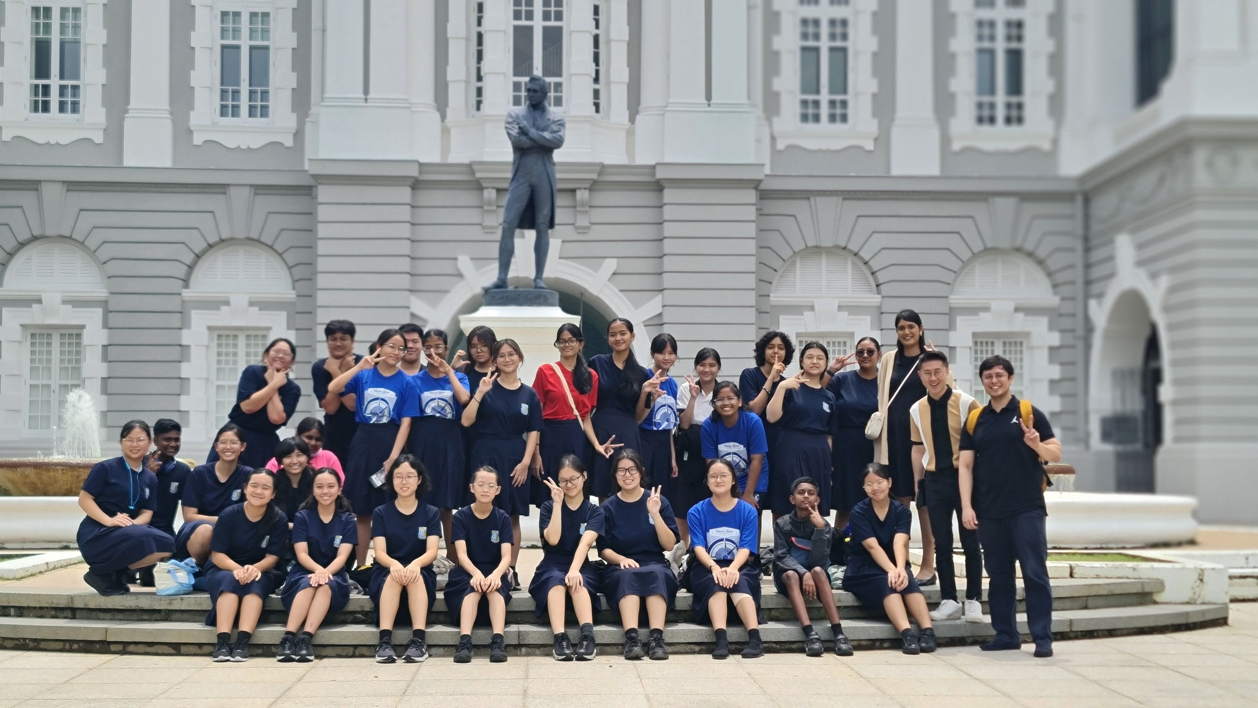 A large group of students in dark blue uniforms pose on steps before a white colonial building with a standing statue.