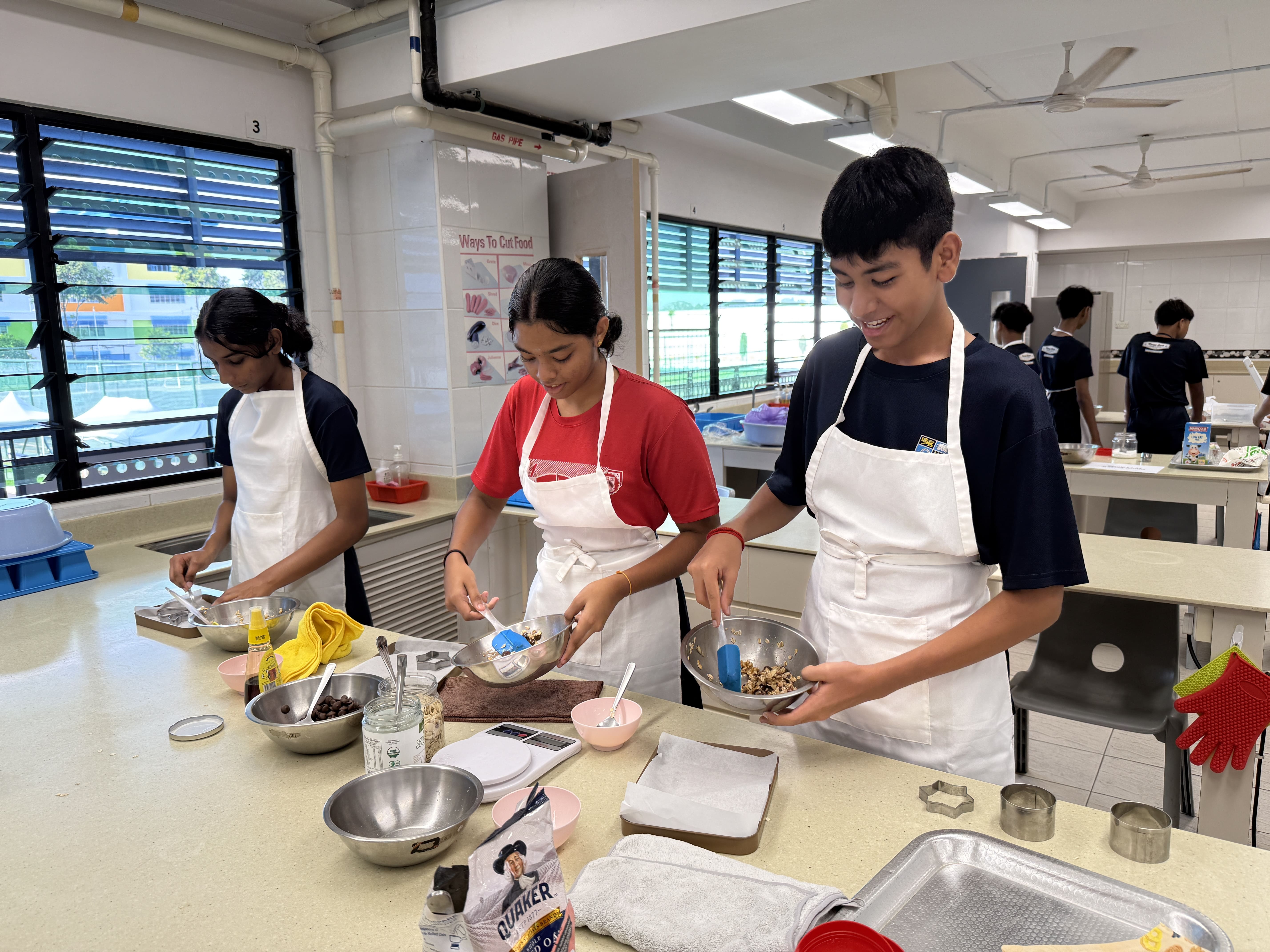 Three students in white aprons mix ingredients in metal bowls at a school cooking classroom counter.