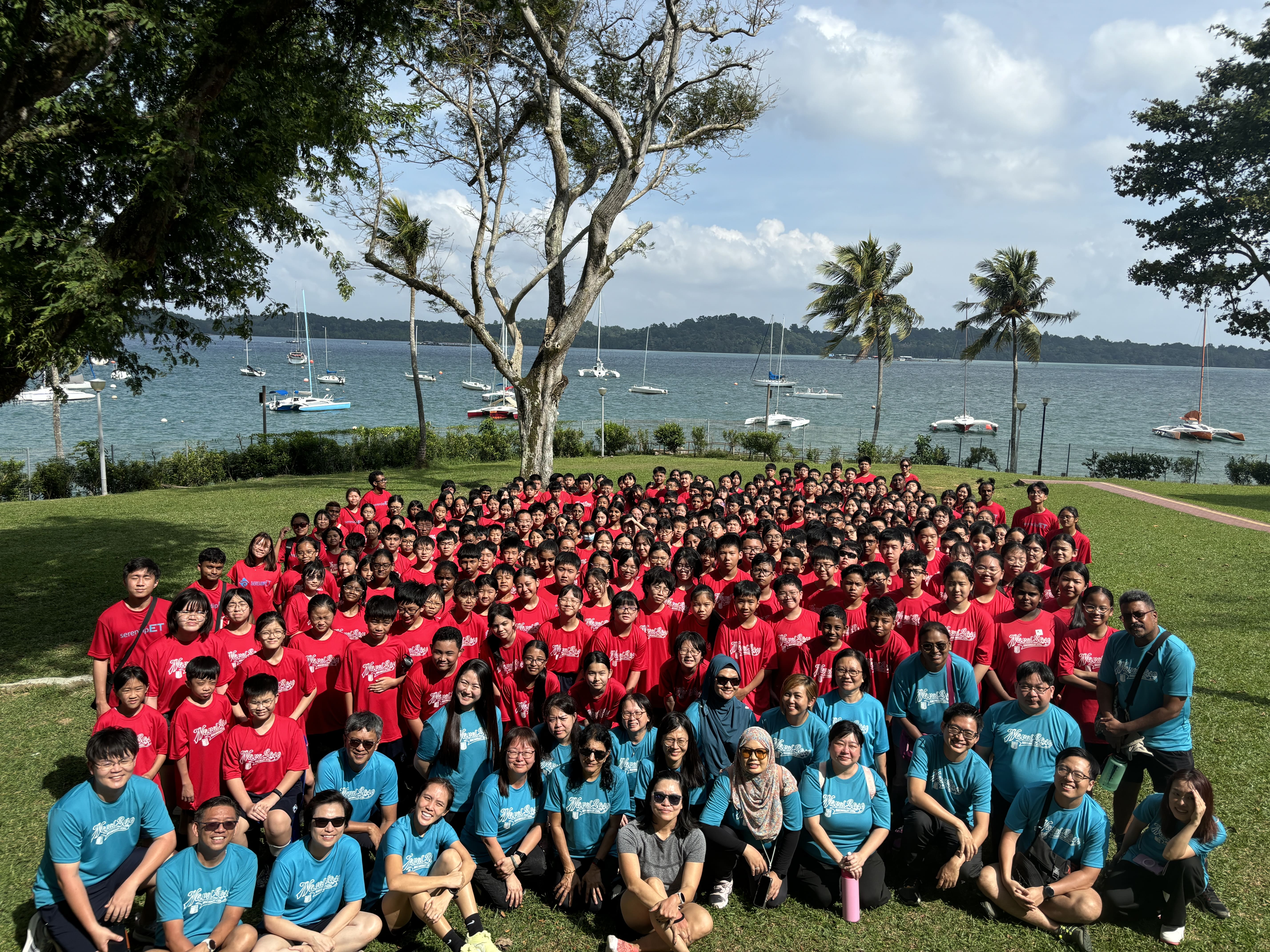 Large group of people in red and teal shirts posing on grass near a waterway with sailboats and palm trees.