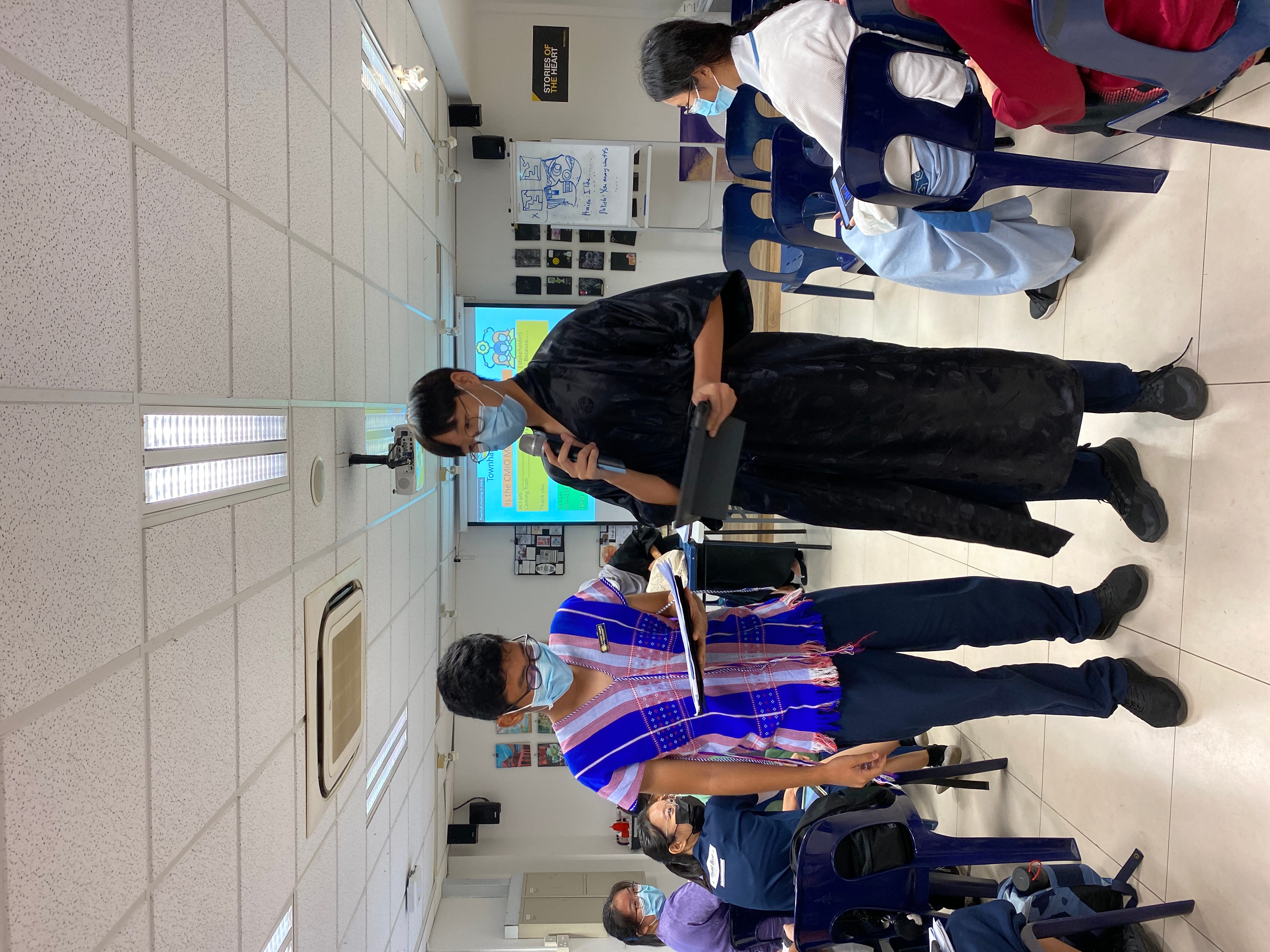 Two masked people stand presenting with a microphone in a classroom, with a projected slideshow and seated audience behind them.
