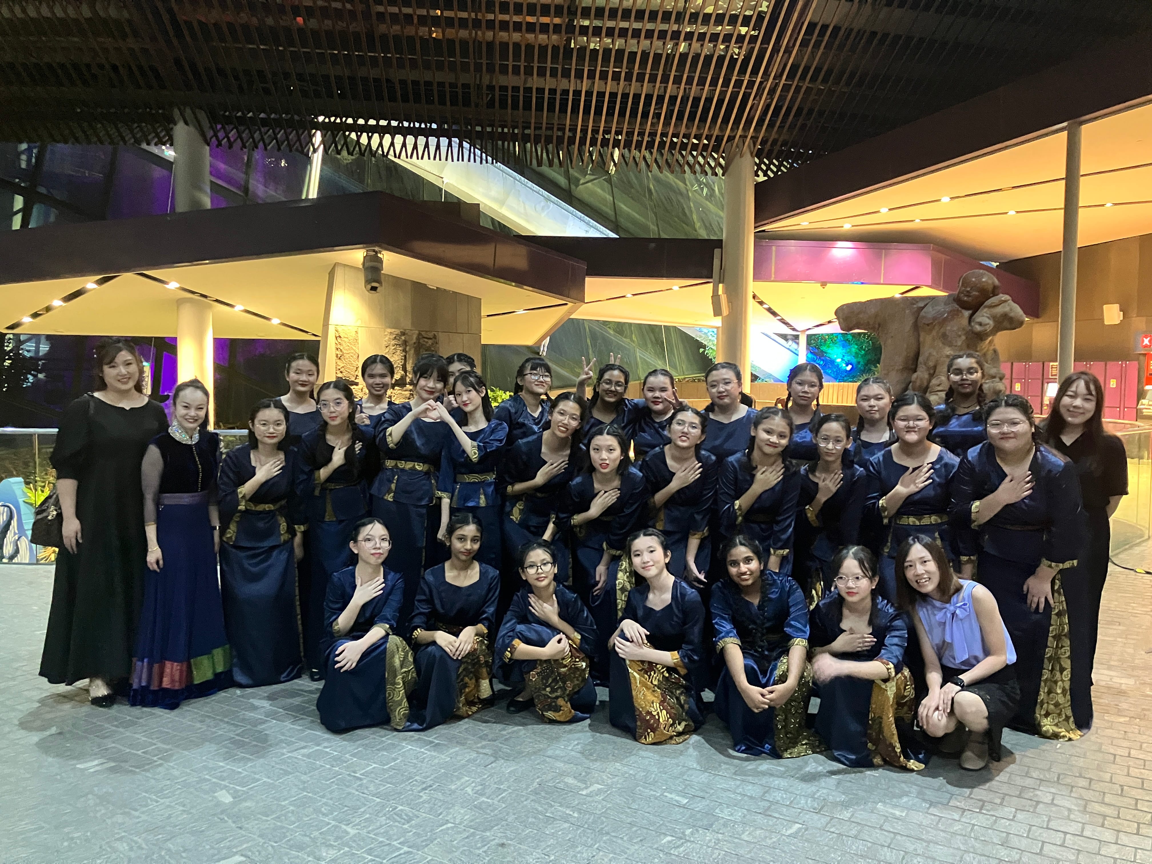 Large group of young women in navy blue and gold outfits posing together outdoors at night near a lit building.
