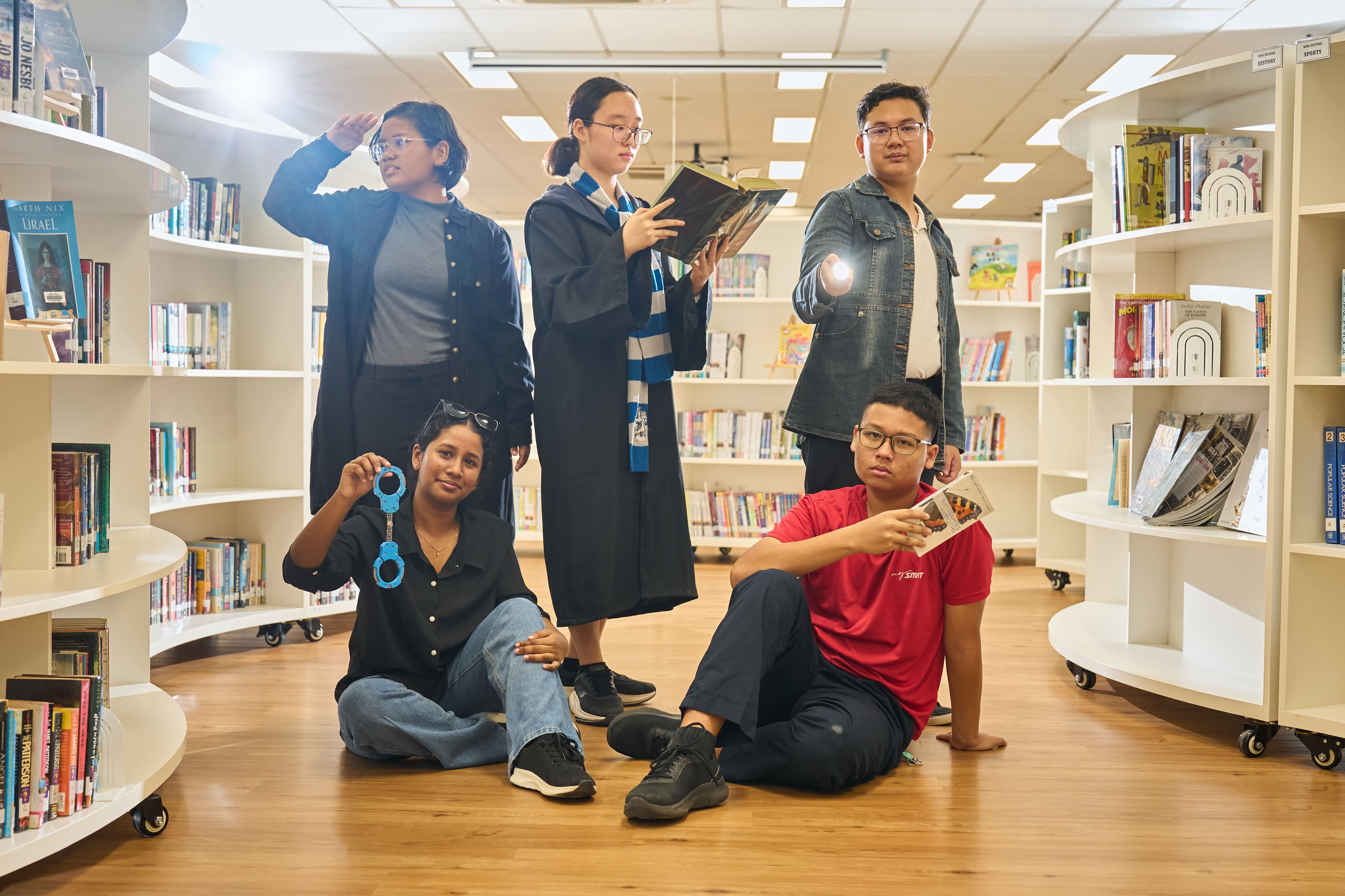 Five students pose dramatically in a library, one in a wizard robe, one holding handcuffs, one shining a flashlight.