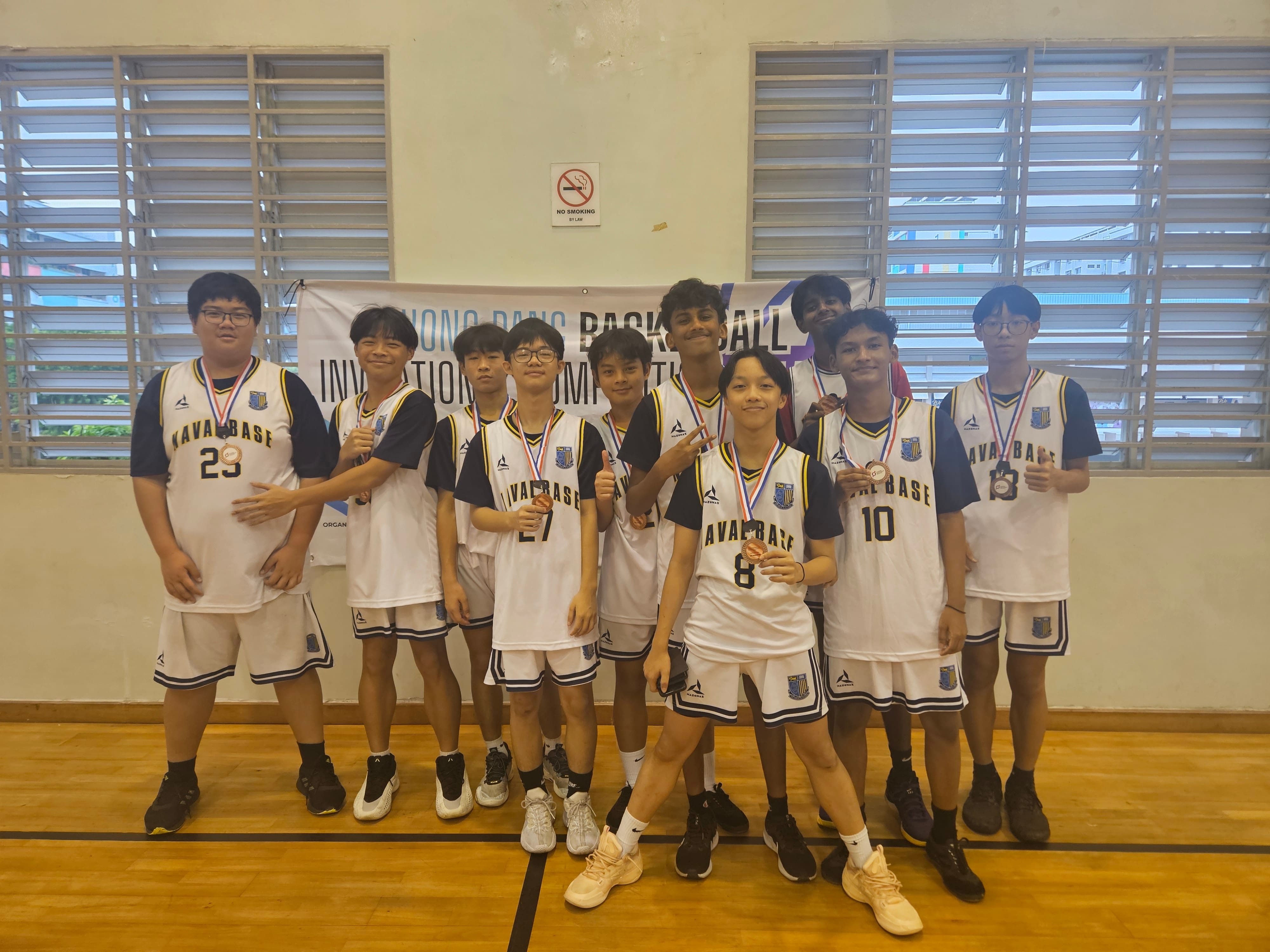 A youth basketball team in white Naval Base jerseys poses with medals in front of a basketball invitational banner.