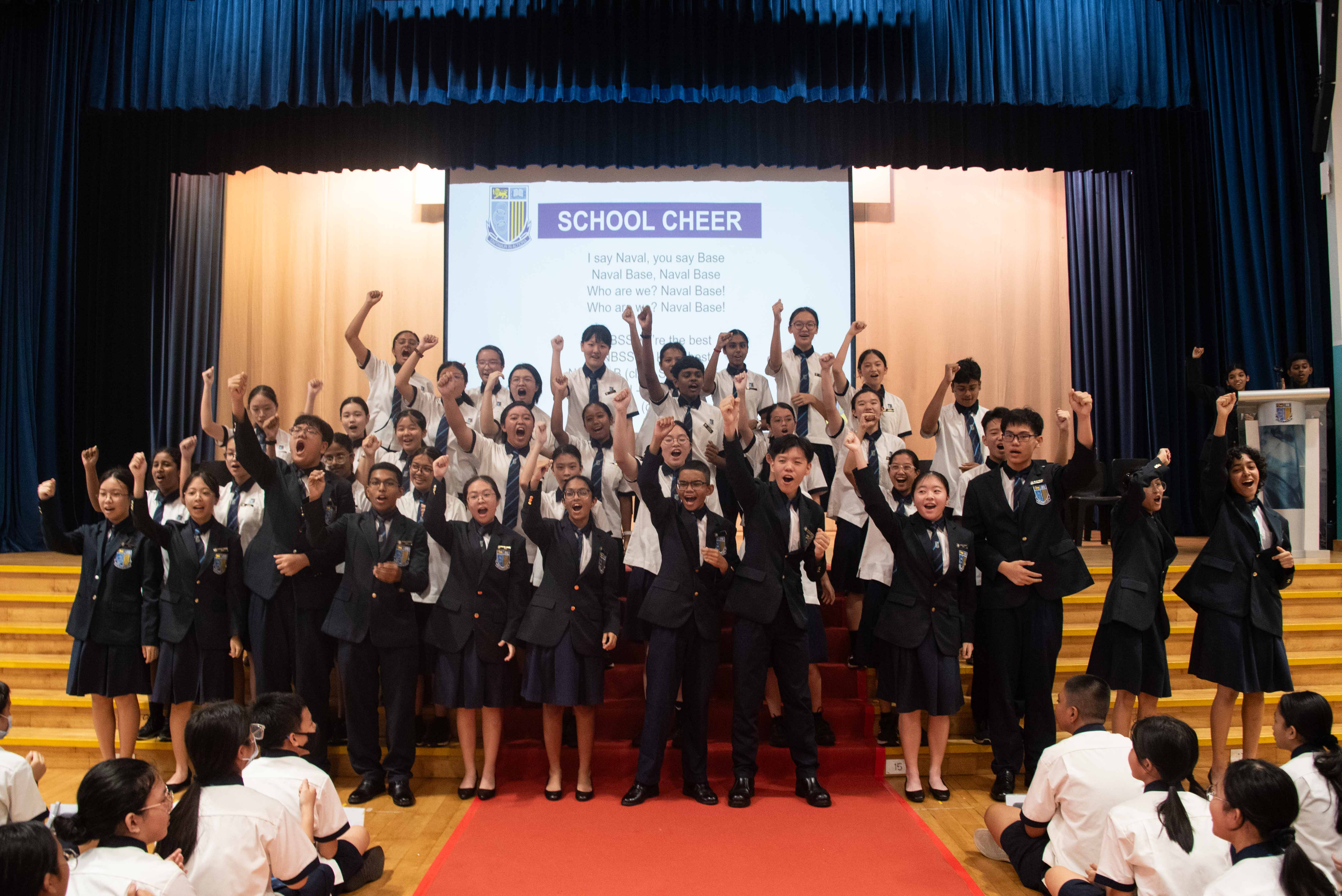 Students in school uniforms cheer with raised fists on a stage in front of a School Cheer slideshow projection.