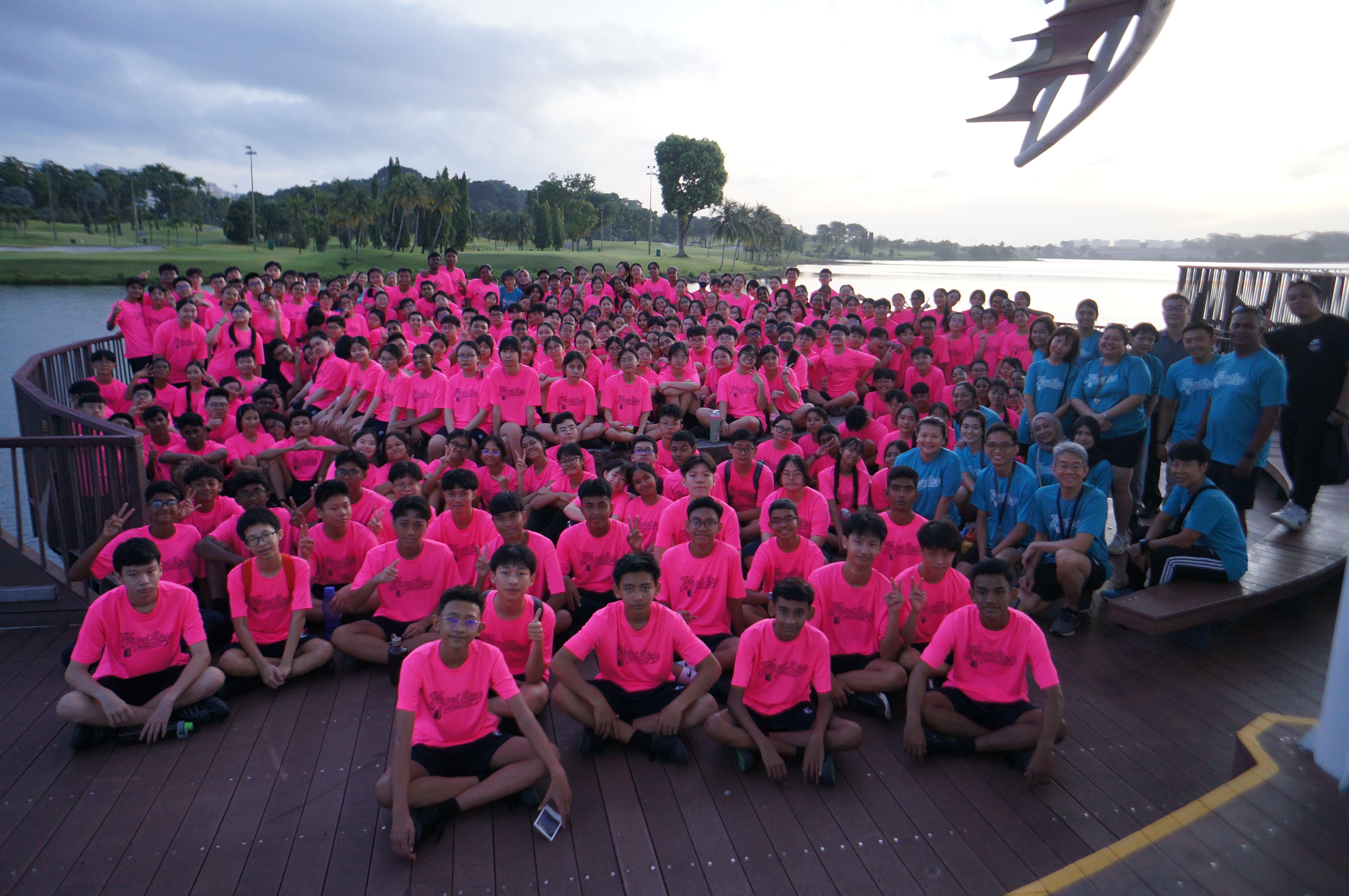 Large group of youth in bright pink shirts posing on a waterfront deck, with a few adults in blue shirts.