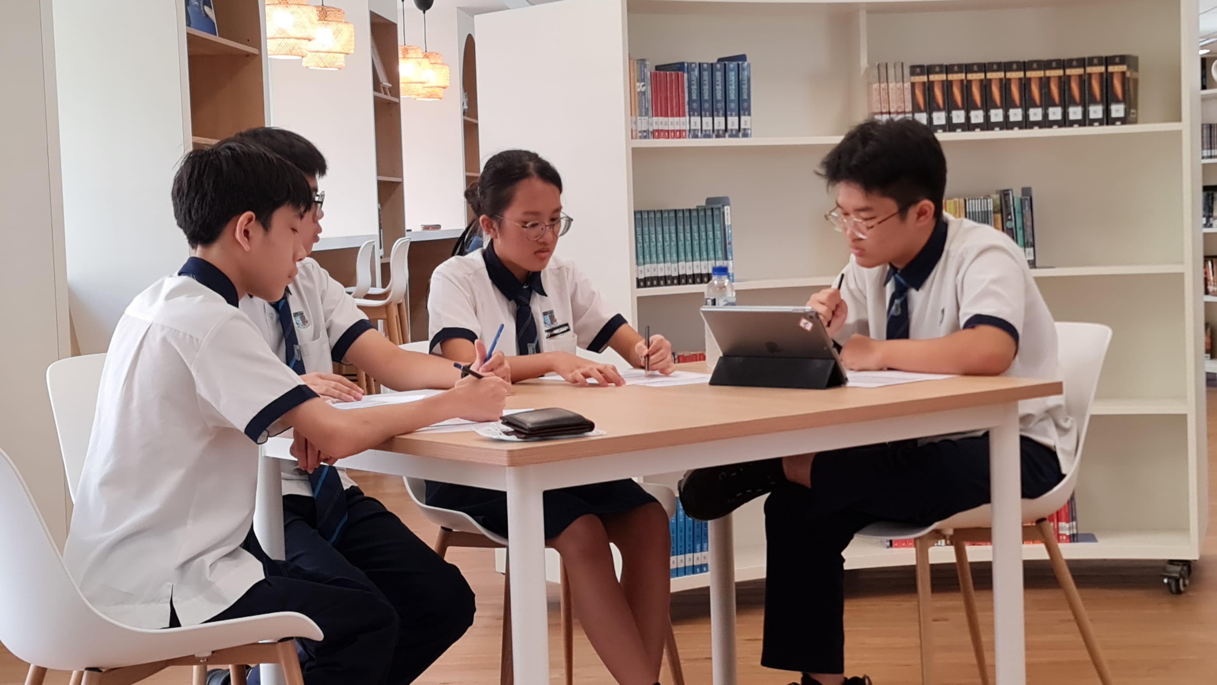 Three uniformed students study together at a table in a library, writing on papers with a tablet nearby.