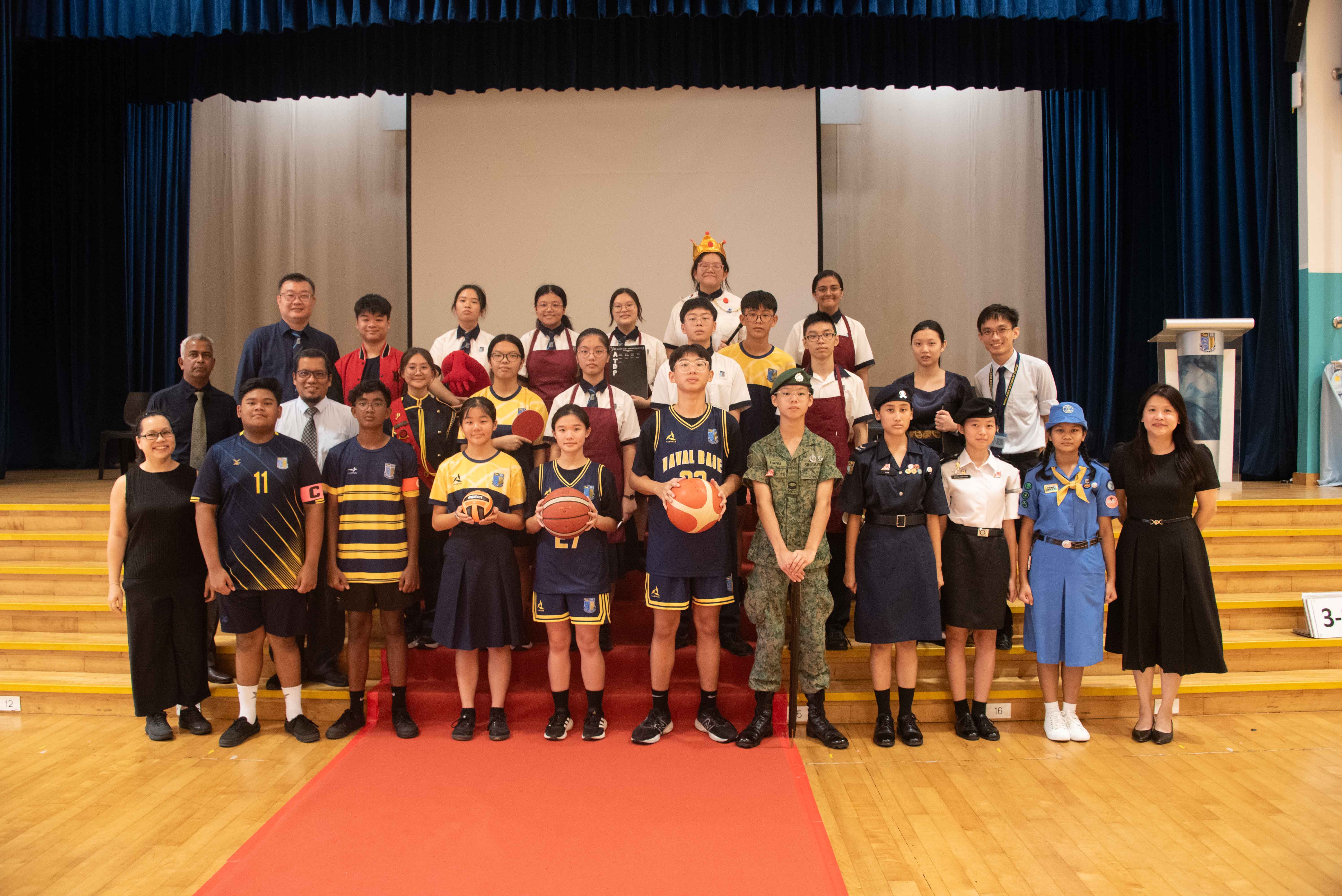 A large group of students and teachers posing on school hall steps, wearing various uniforms including sports, military, scout, and cooking attire.