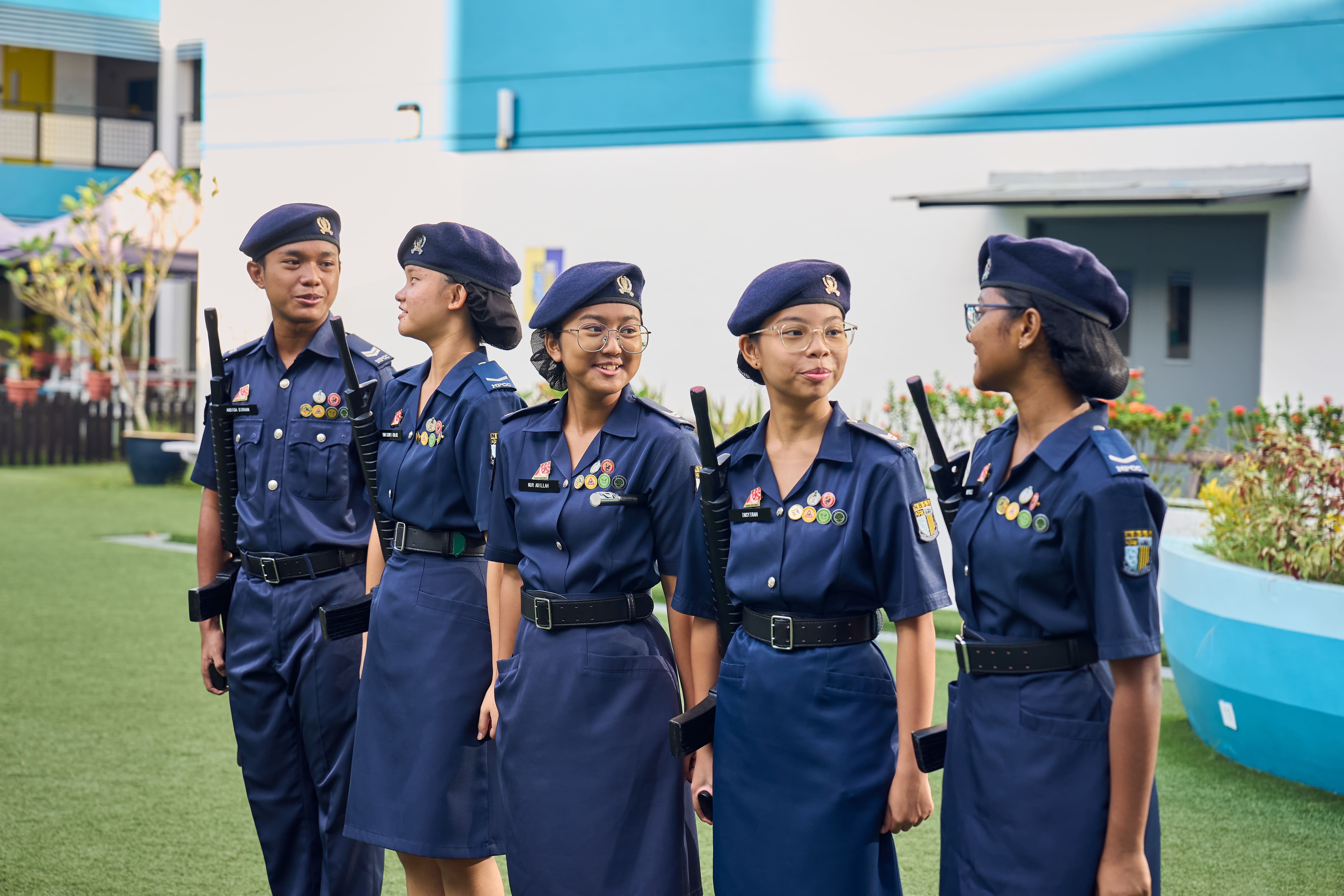 Five young cadets in navy blue uniforms and berets with badges stand outdoors on grass, smiling and talking.
