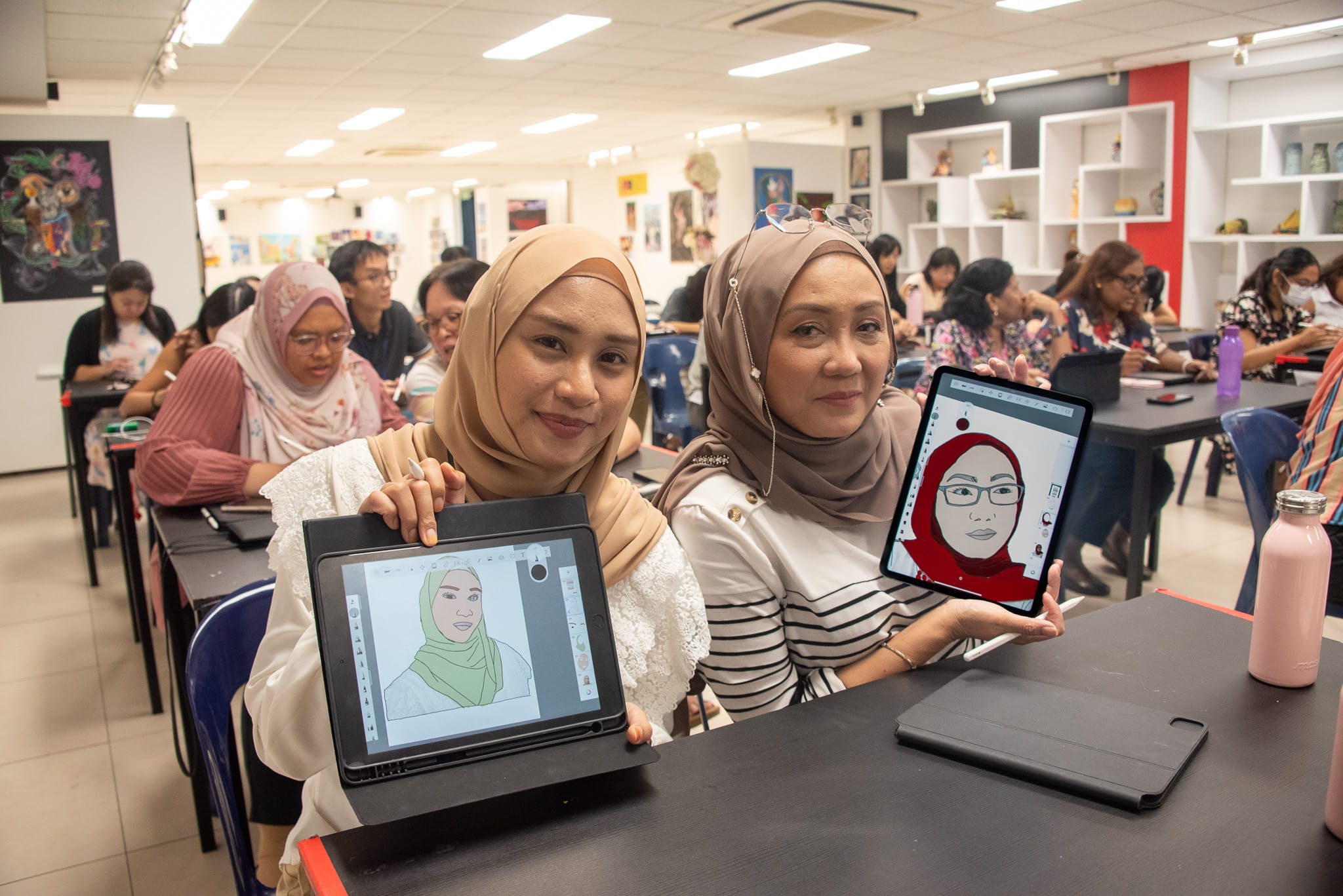 Two women wearing hijabs hold up tablets showing digital portrait drawings of themselves in a busy classroom.