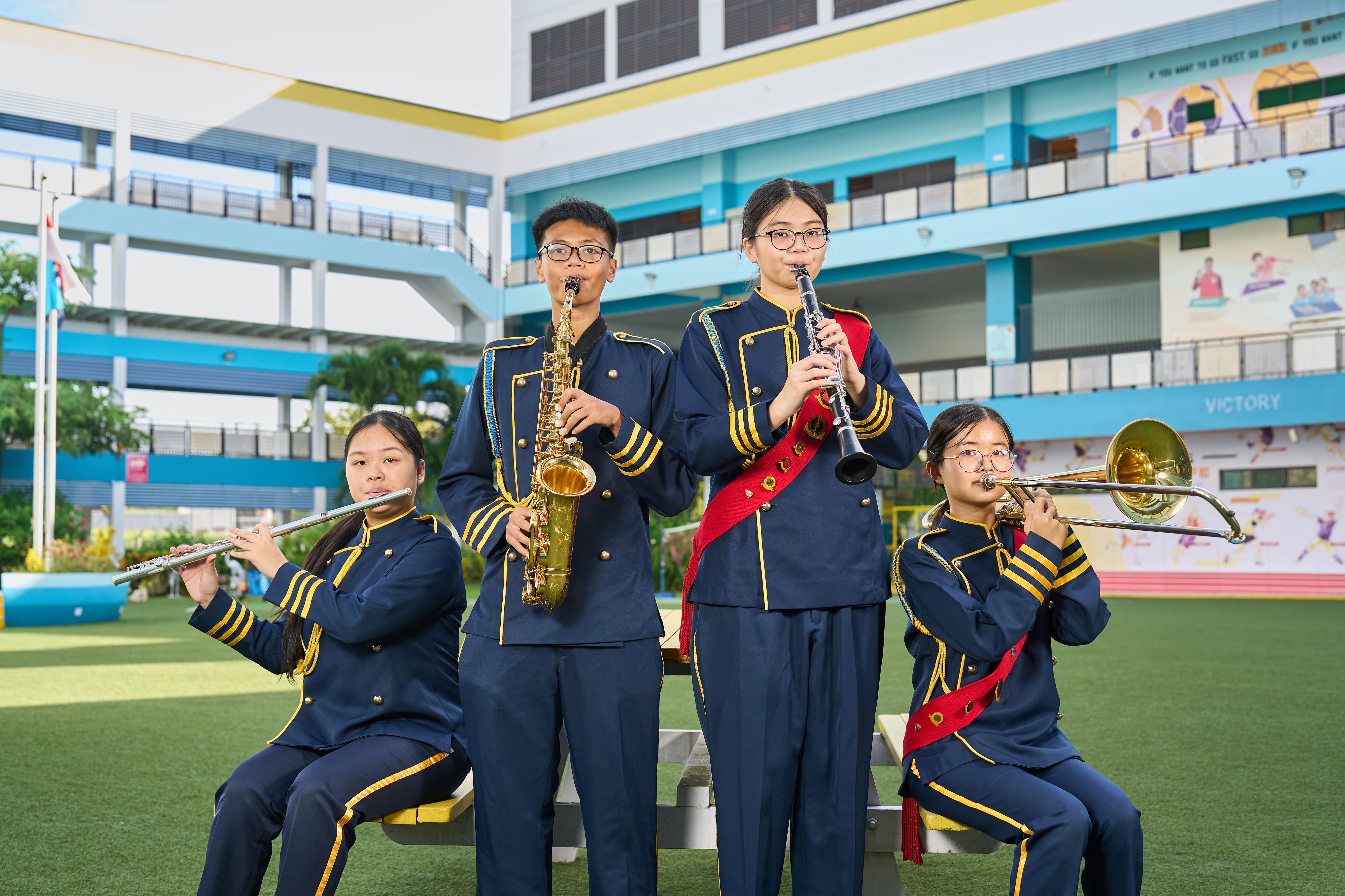 Four students in navy marching band uniforms play flute, saxophone, clarinet, and trombone outdoors at a school.
