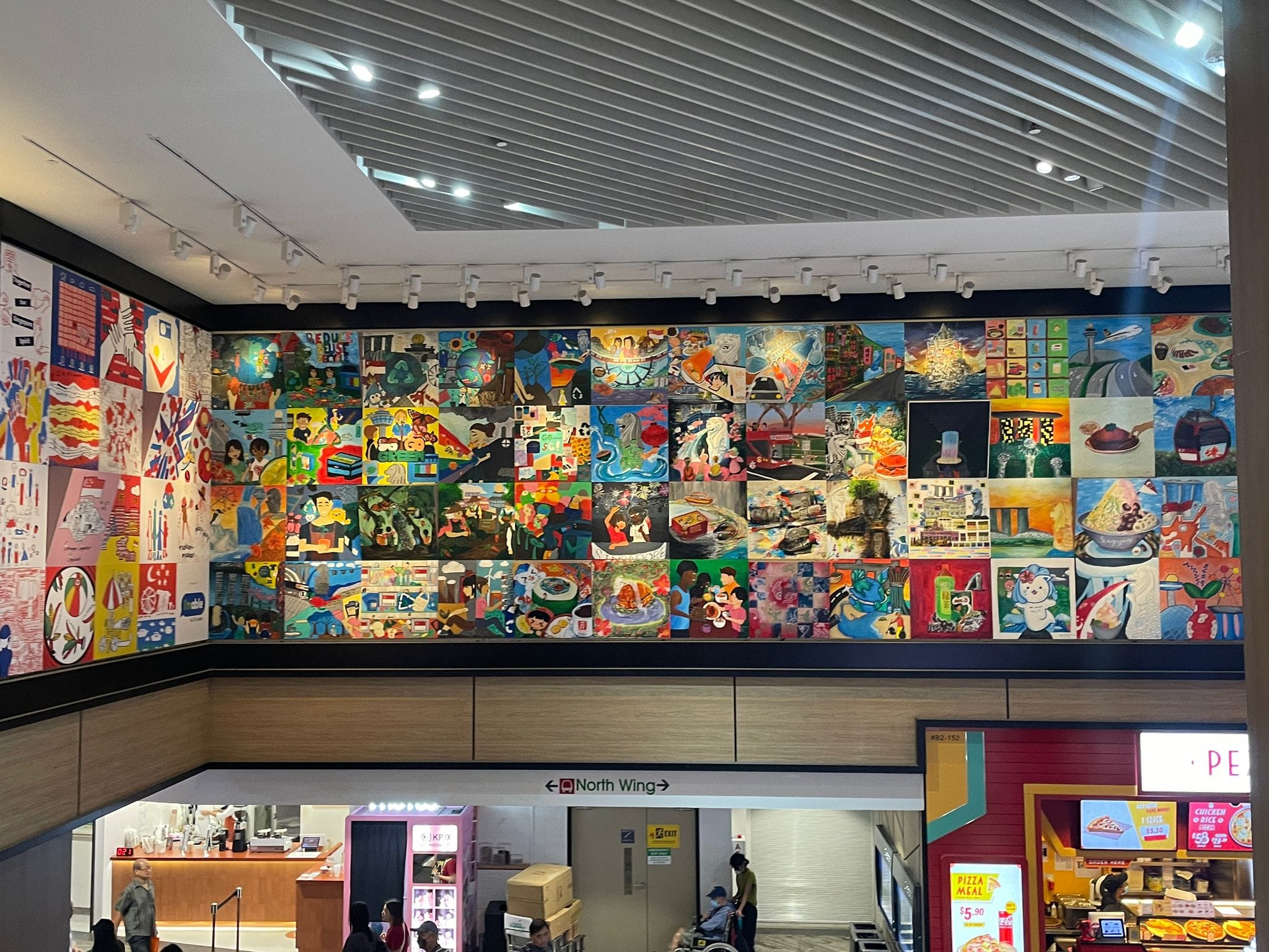 A large colorful mural made of many painted panels covers an upper wall inside a shopping mall above food stalls.