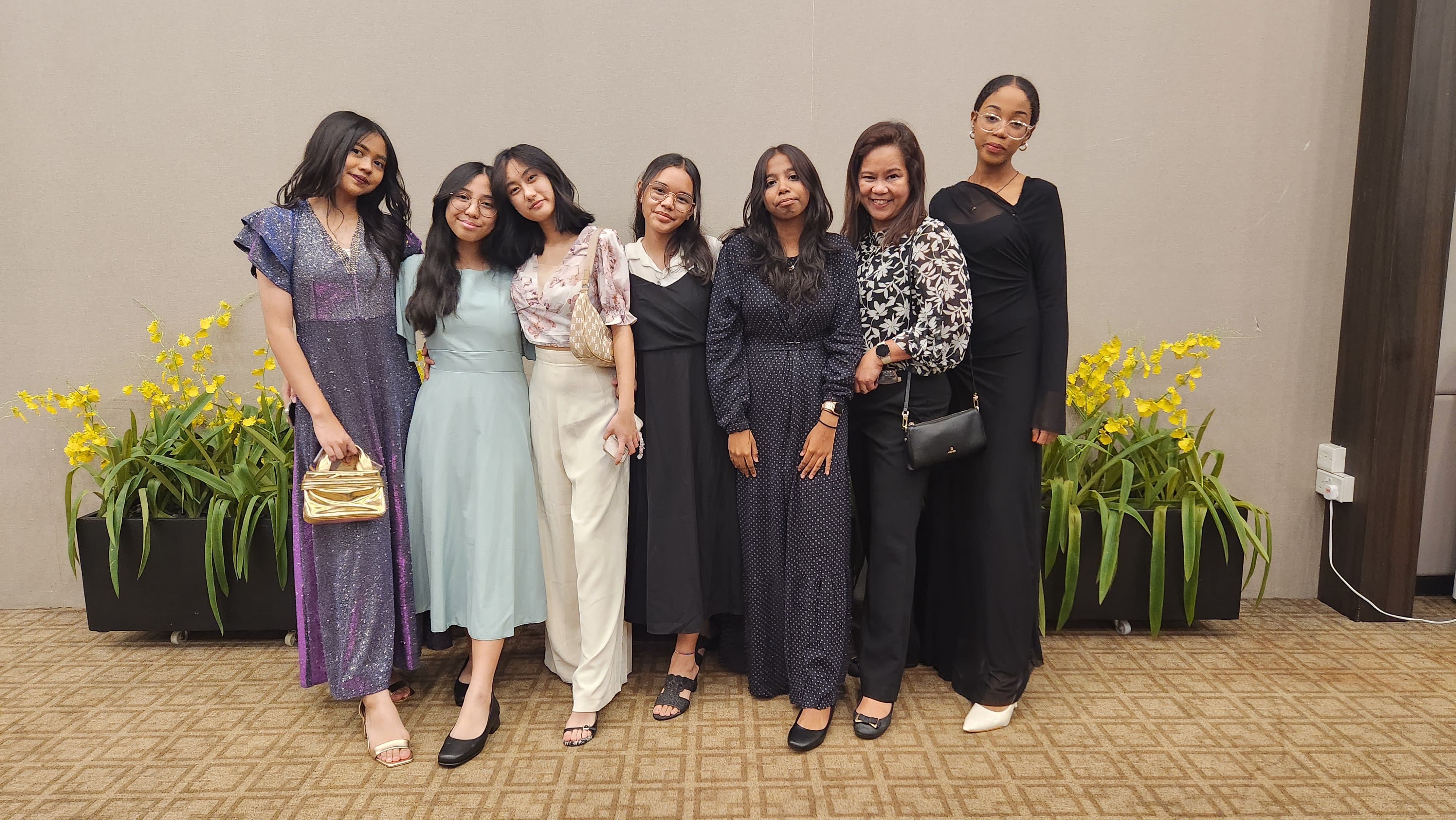 Seven women in formal attire pose together smiling, with yellow flowering plants behind them indoors.