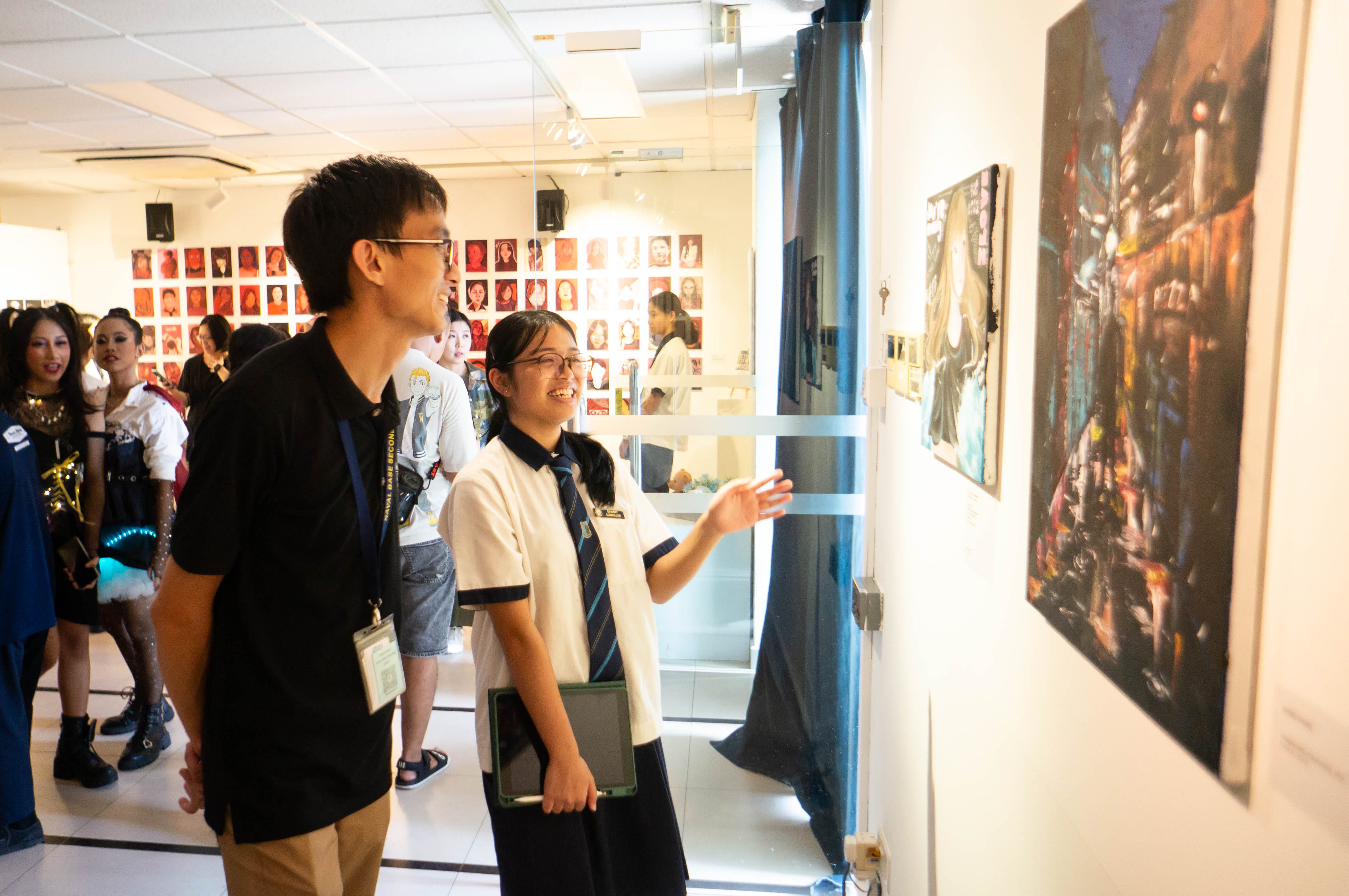 Two smiling students view paintings on a gallery wall, with portrait artworks and a crowd visible in the background.